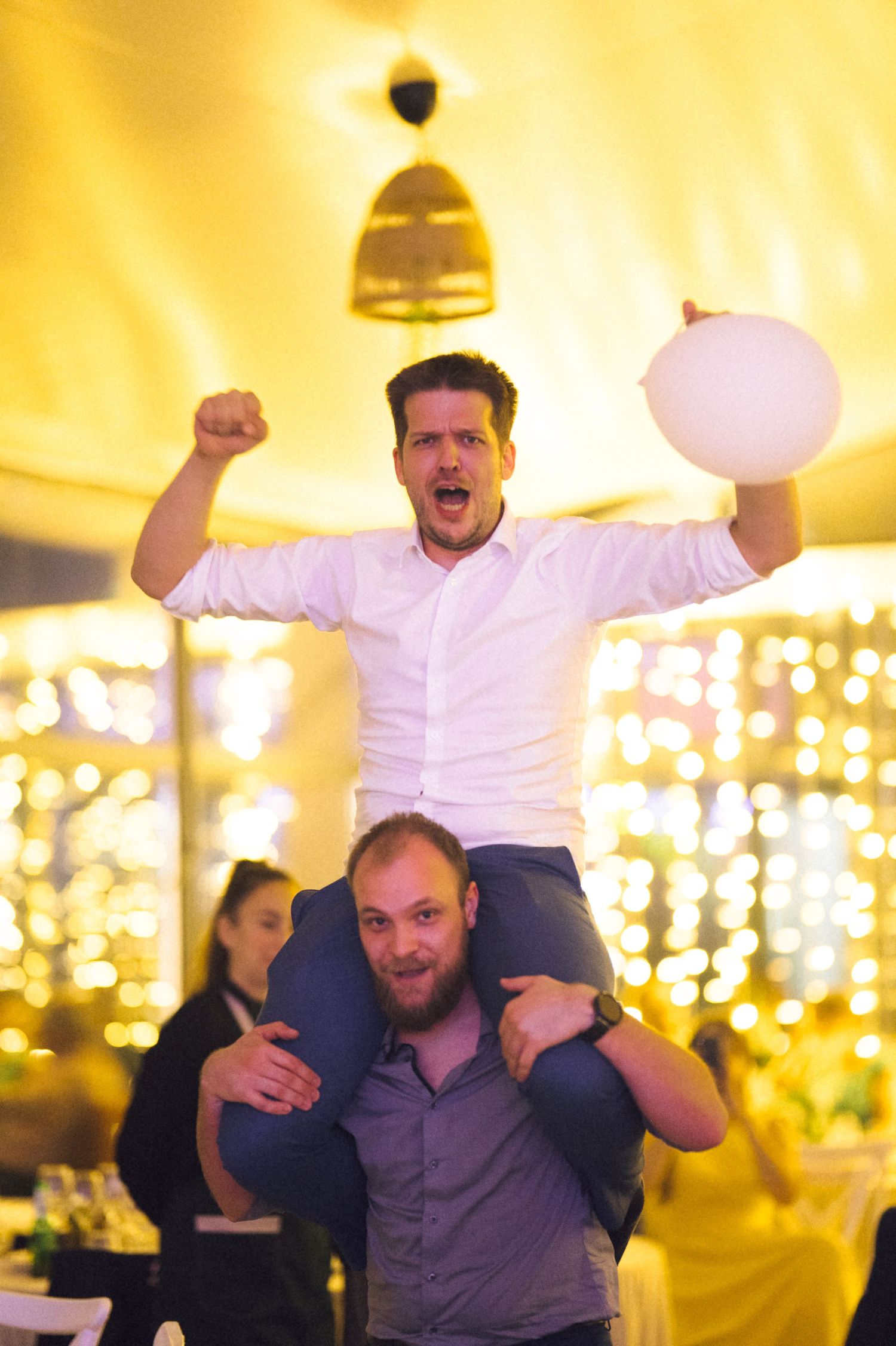 Man in white shirt celebrating on another man's shoulders, holding balloon at festive event with warm bokeh lights.