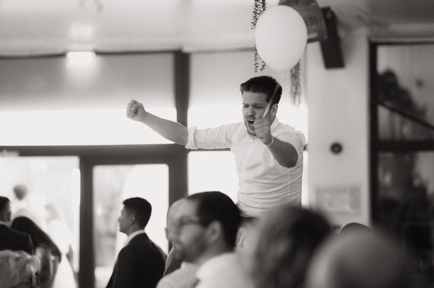 Man celebrating with raised fist at indoor event, black and white photo with balloon and seated guests.