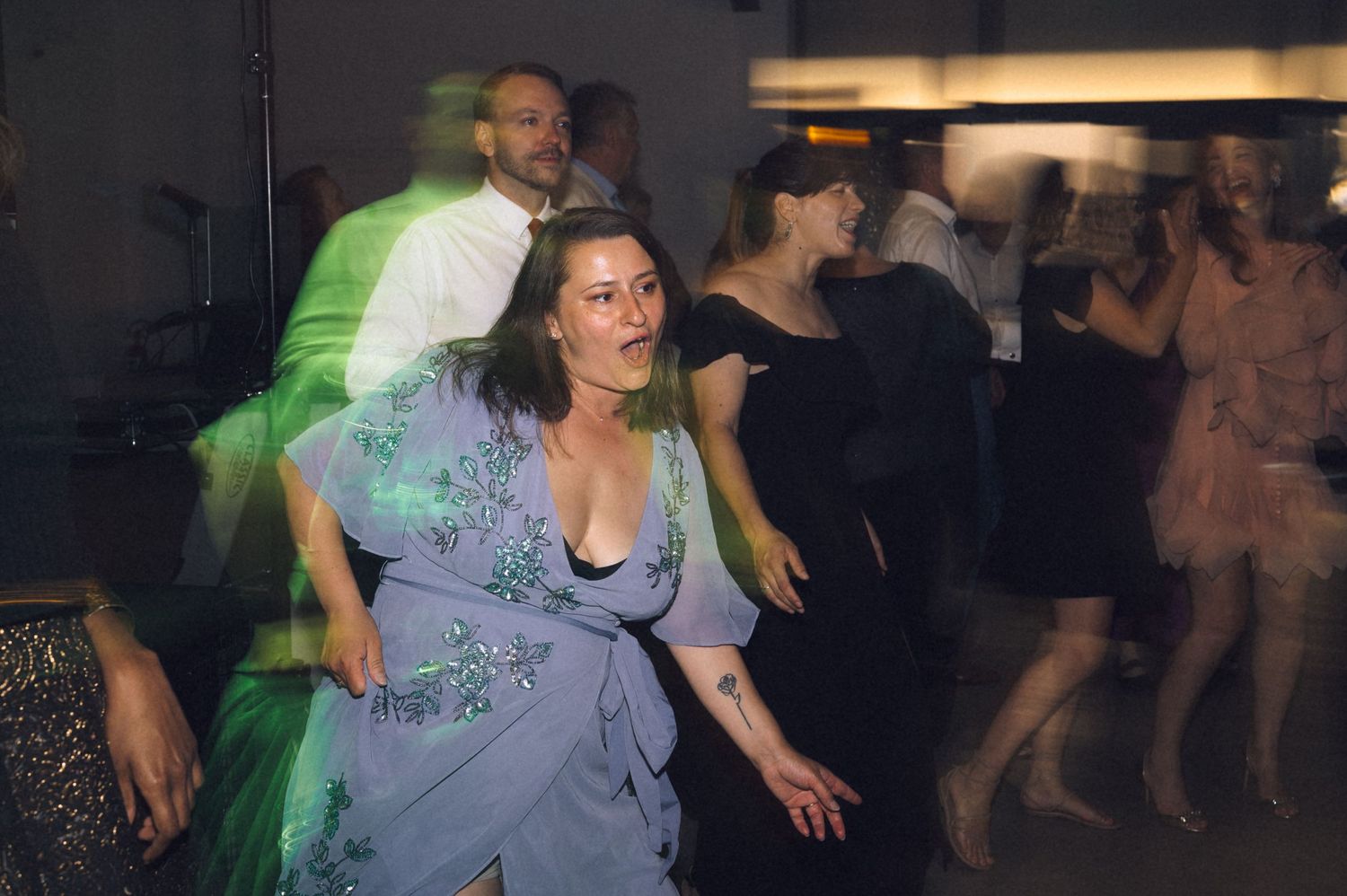 Guests dancing energetically at a formal event, woman in blue floral gown in foreground, motion blur effect.
