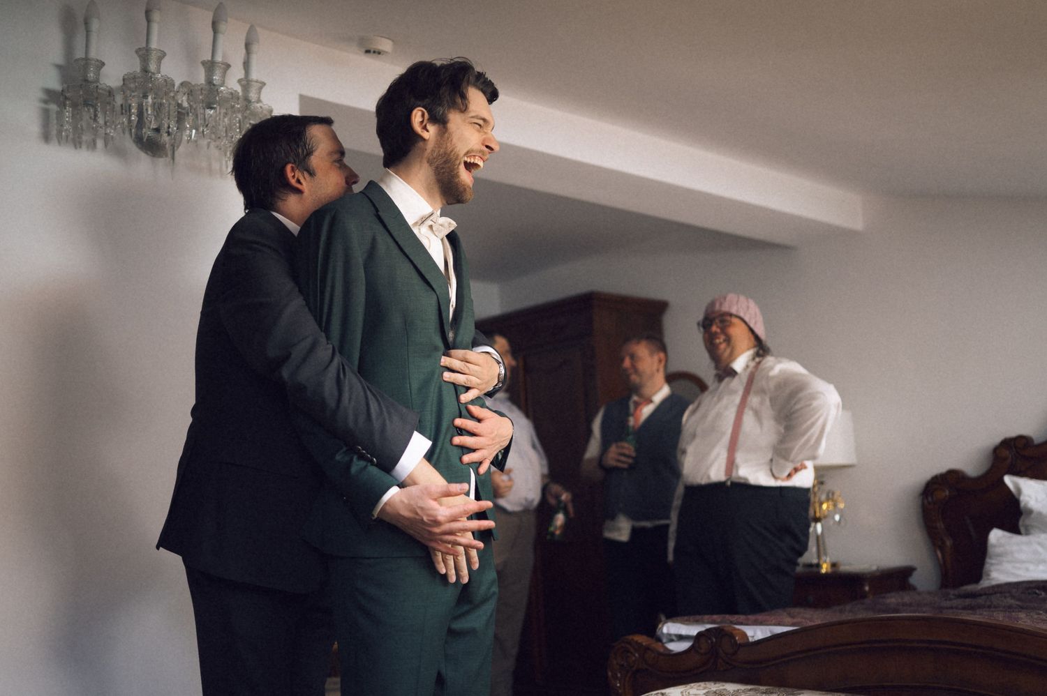 Men in formal suits laughing and celebrating together indoors at a wedding preparation gathering.