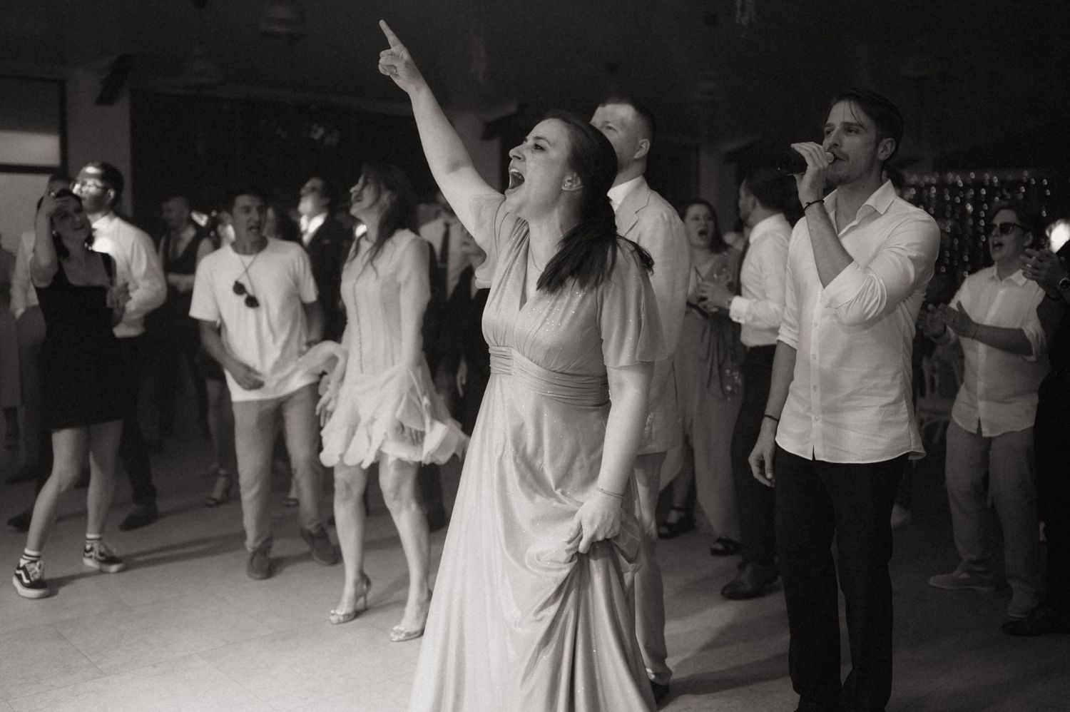 Black and white photo of people dancing at a party, woman in dress pointing up joyfully.