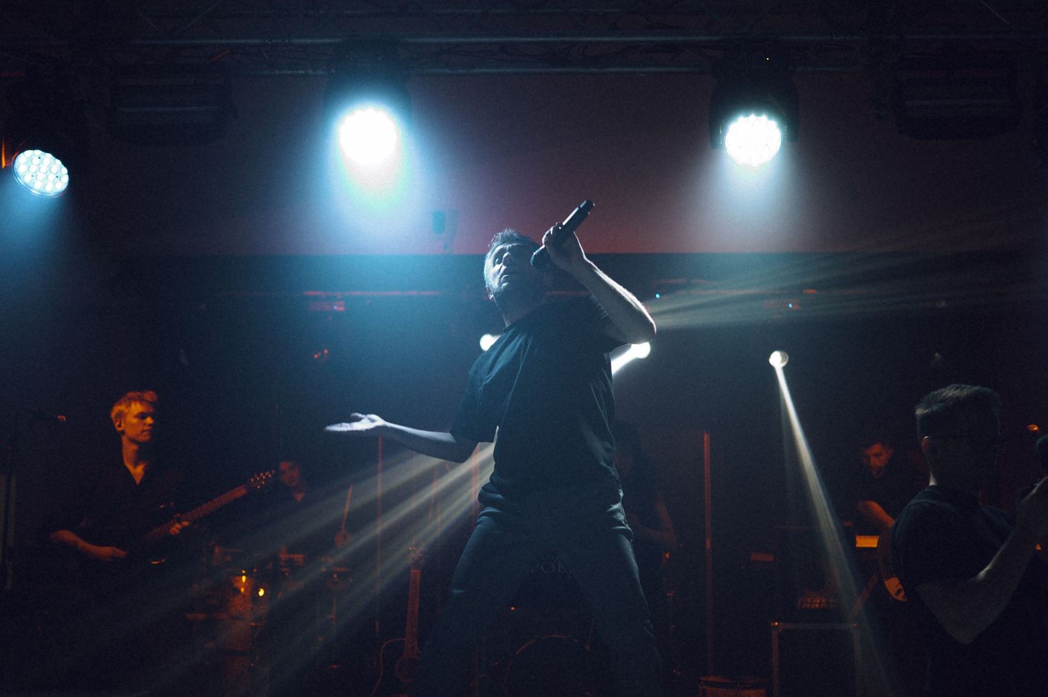 Singer performing on stage under blue and white spotlights in a dark concert venue with band members in background.