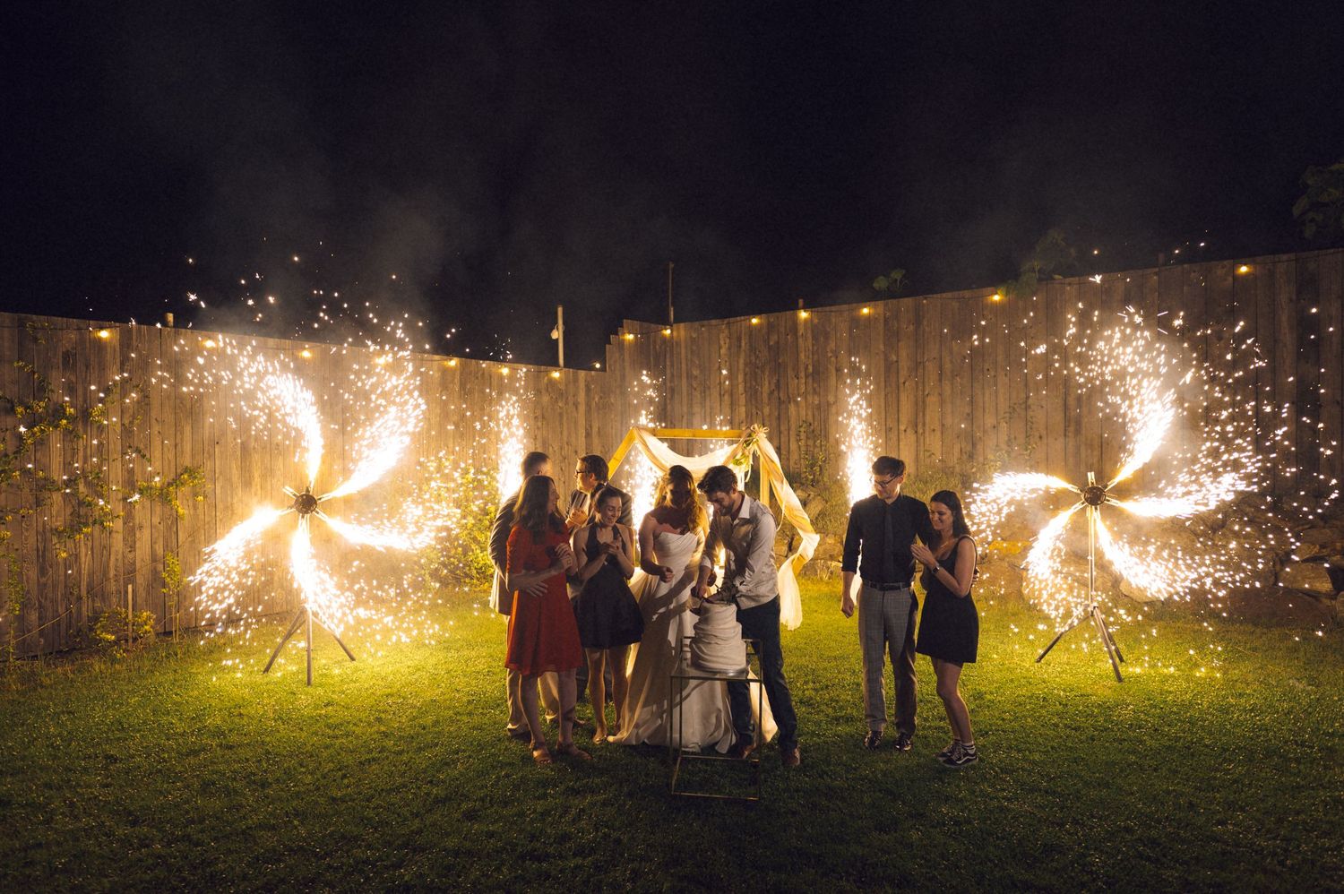 Wedding couple kissing surrounded by guests with spinning sparklers and string lights at night outdoor celebration.