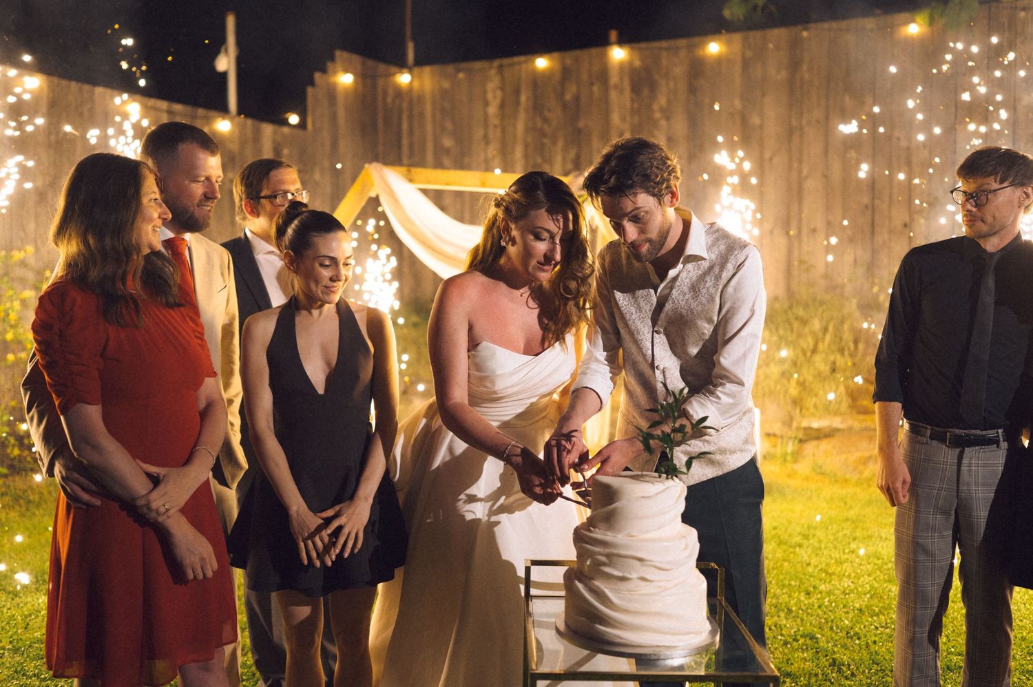 Bride and groom cutting wedding cake outdoors at night with sparklers and guests gathered around at a backyard celebration.