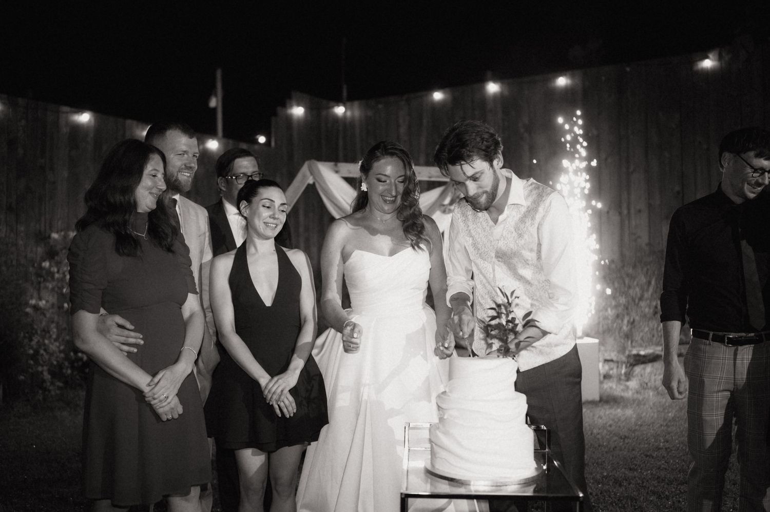 Black and white photo of bride and groom cutting wedding cake surrounded by smiling guests with string lights in background.