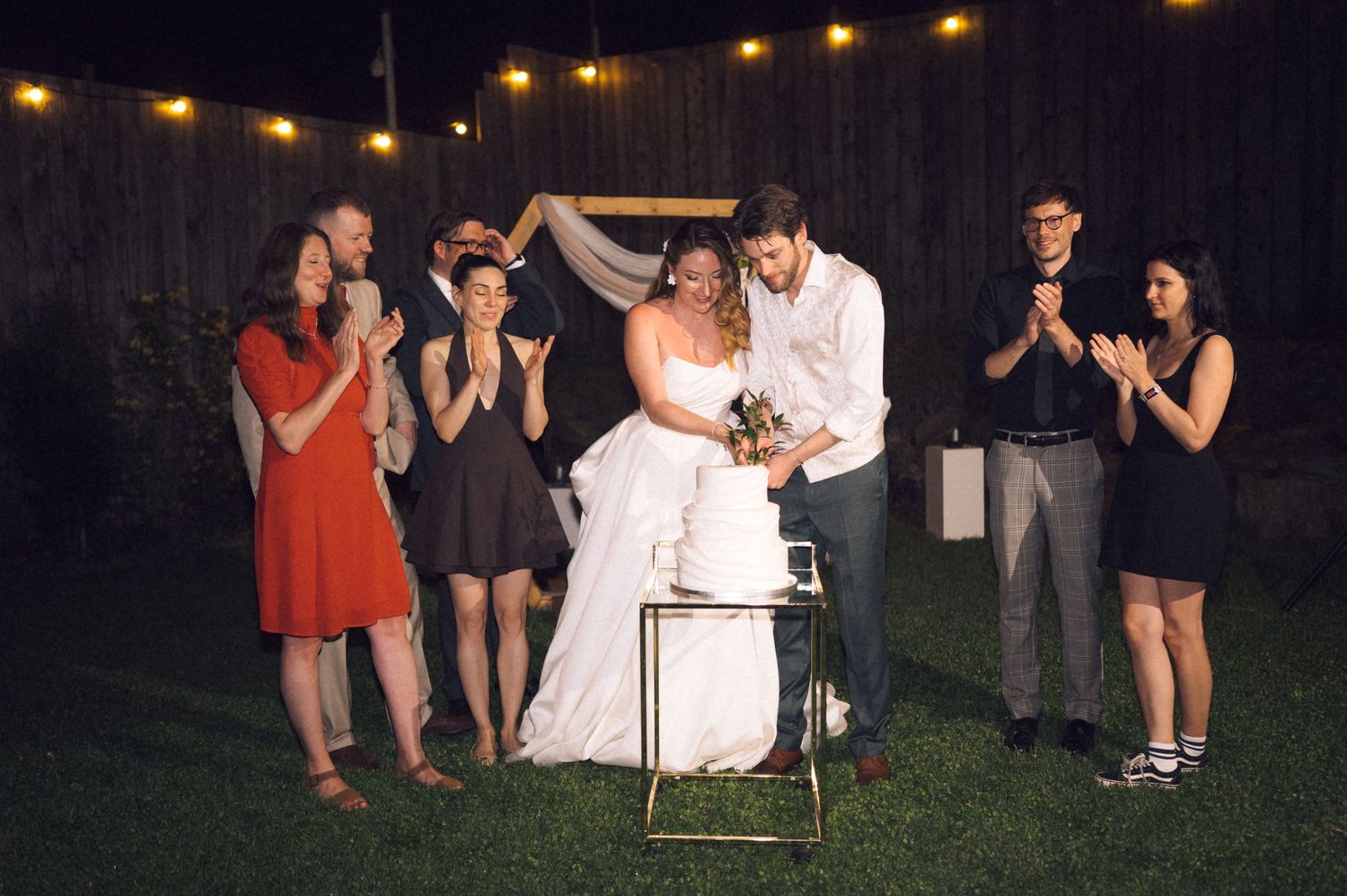 Newlywed couple shares first kiss at outdoor backyard wedding ceremony at night, surrounded by applauding guests under string lights.