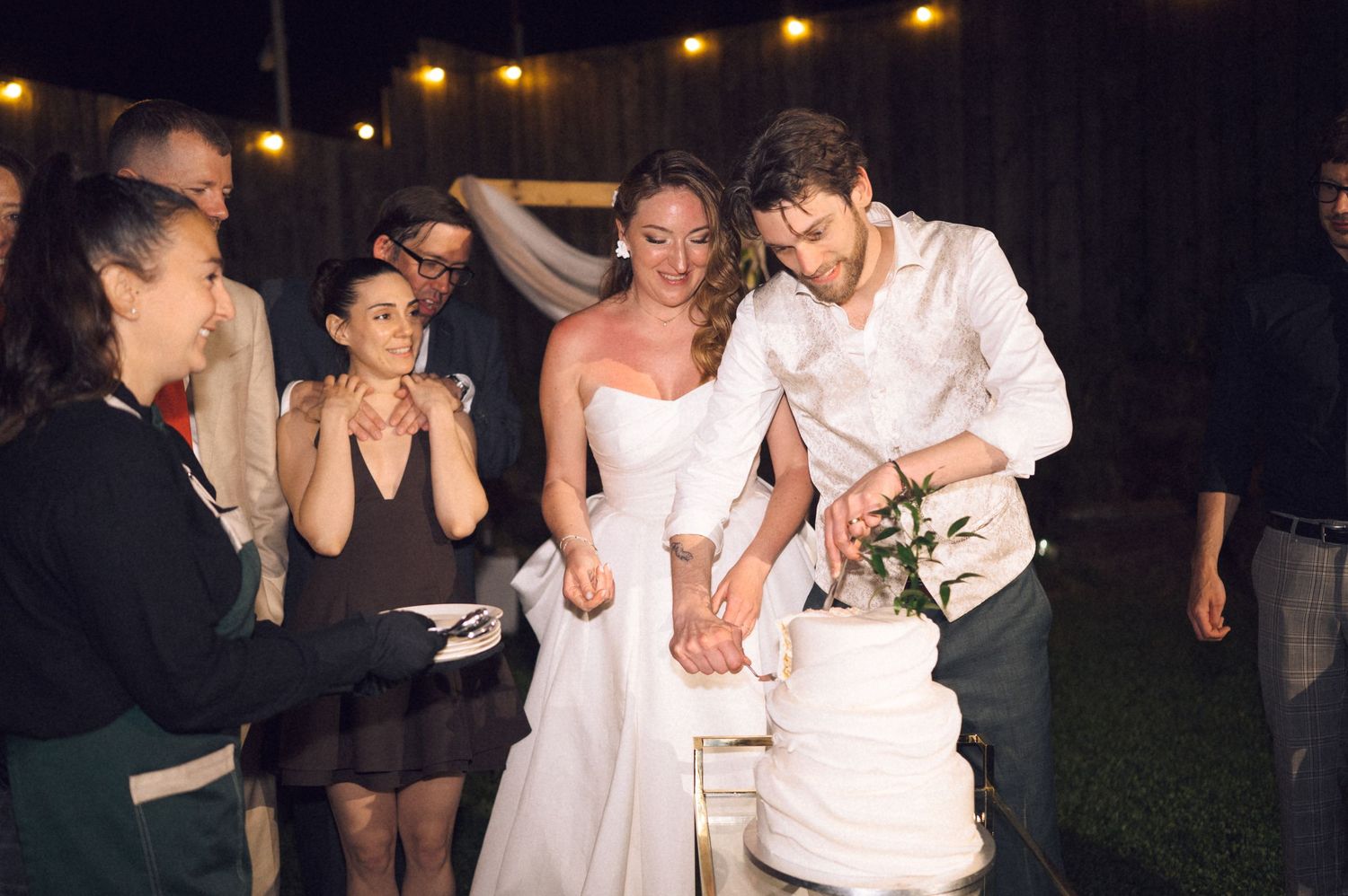 Bride and groom cutting wedding cake together outdoors at night surrounded by smiling guests under string lights.
