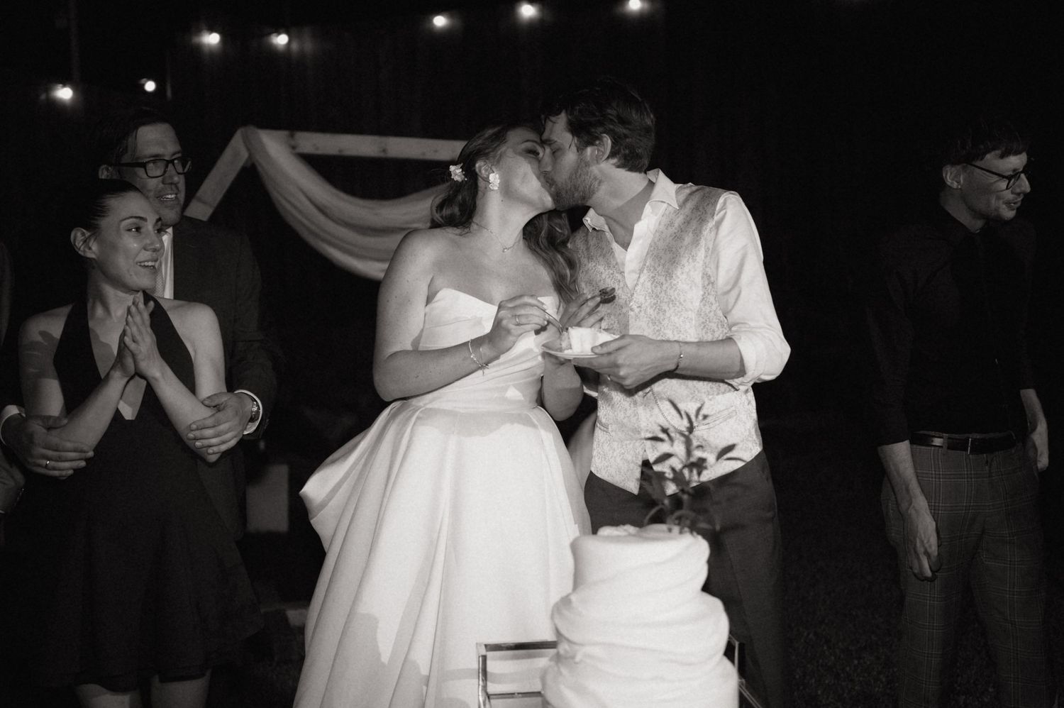 Bride and groom share a kiss while cutting their wedding cake surrounded by guests.