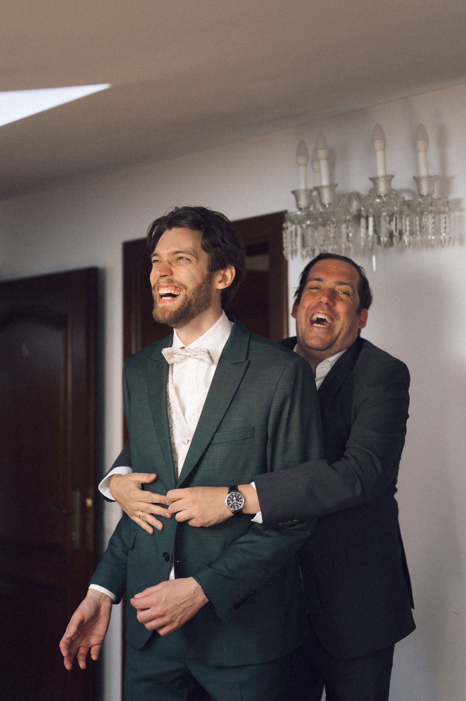 Two men in suits laughing joyfully, one in green with bow tie, other in black, sharing a fun moment indoors.