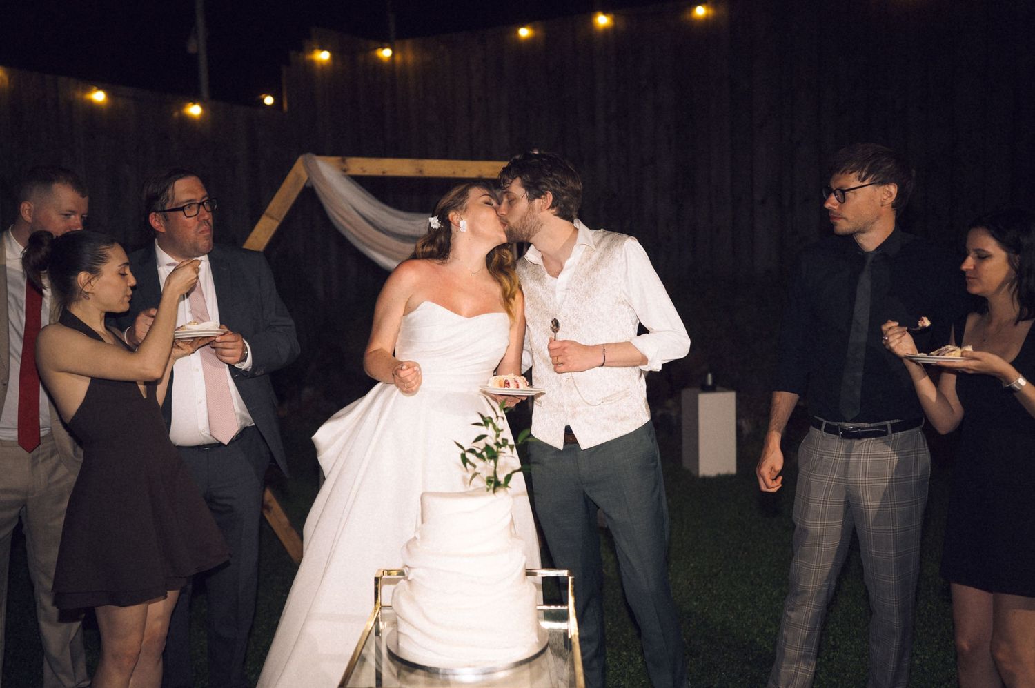 Bride and groom share a kiss beside their wedding cake at an outdoor evening reception with guests gathered around.