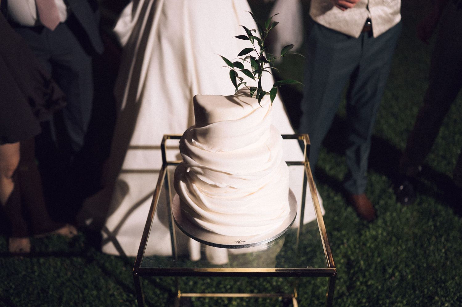 Elegant three-tier white wedding cake with greenery topper on gold stand, surrounded by guests on grass at night.