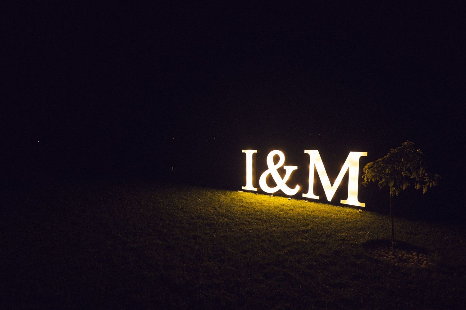 Illuminated I&M letter sign glowing in golden light on grass at night outdoor wedding decor.