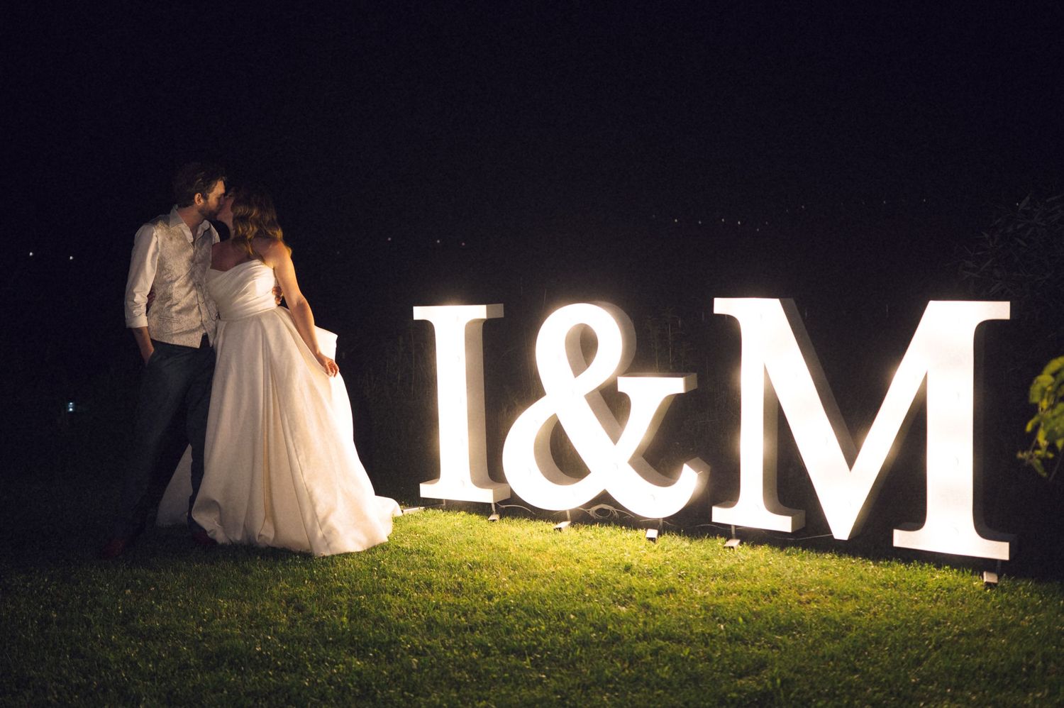 Bride and groom kissing beside illuminated I&M monogram letters at night wedding celebration outdoors.