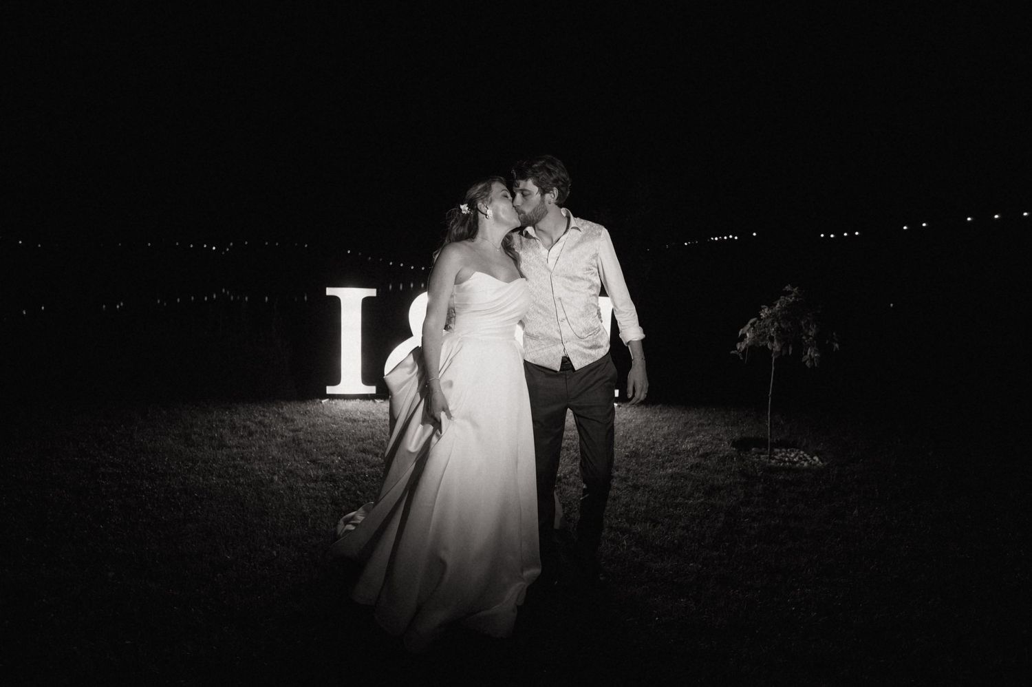 Black and white wedding photo of couple kissing at night near illuminated LOVE letters outdoors.