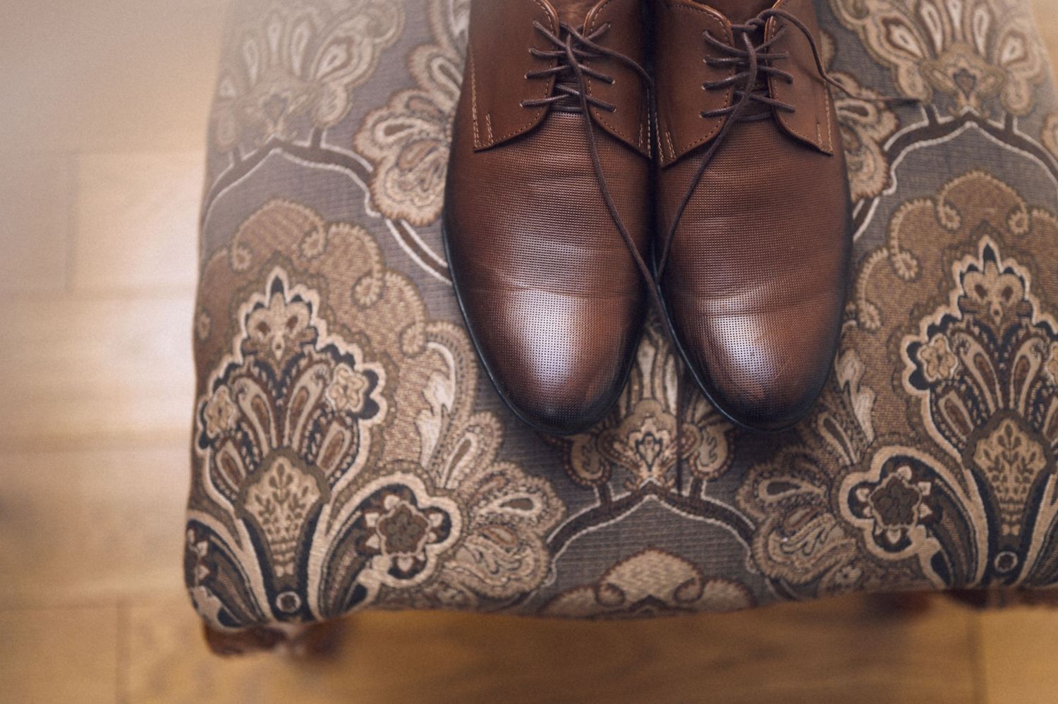 Brown leather lace-up dress shoes displayed on an ornate paisley upholstered ottoman on hardwood floor.