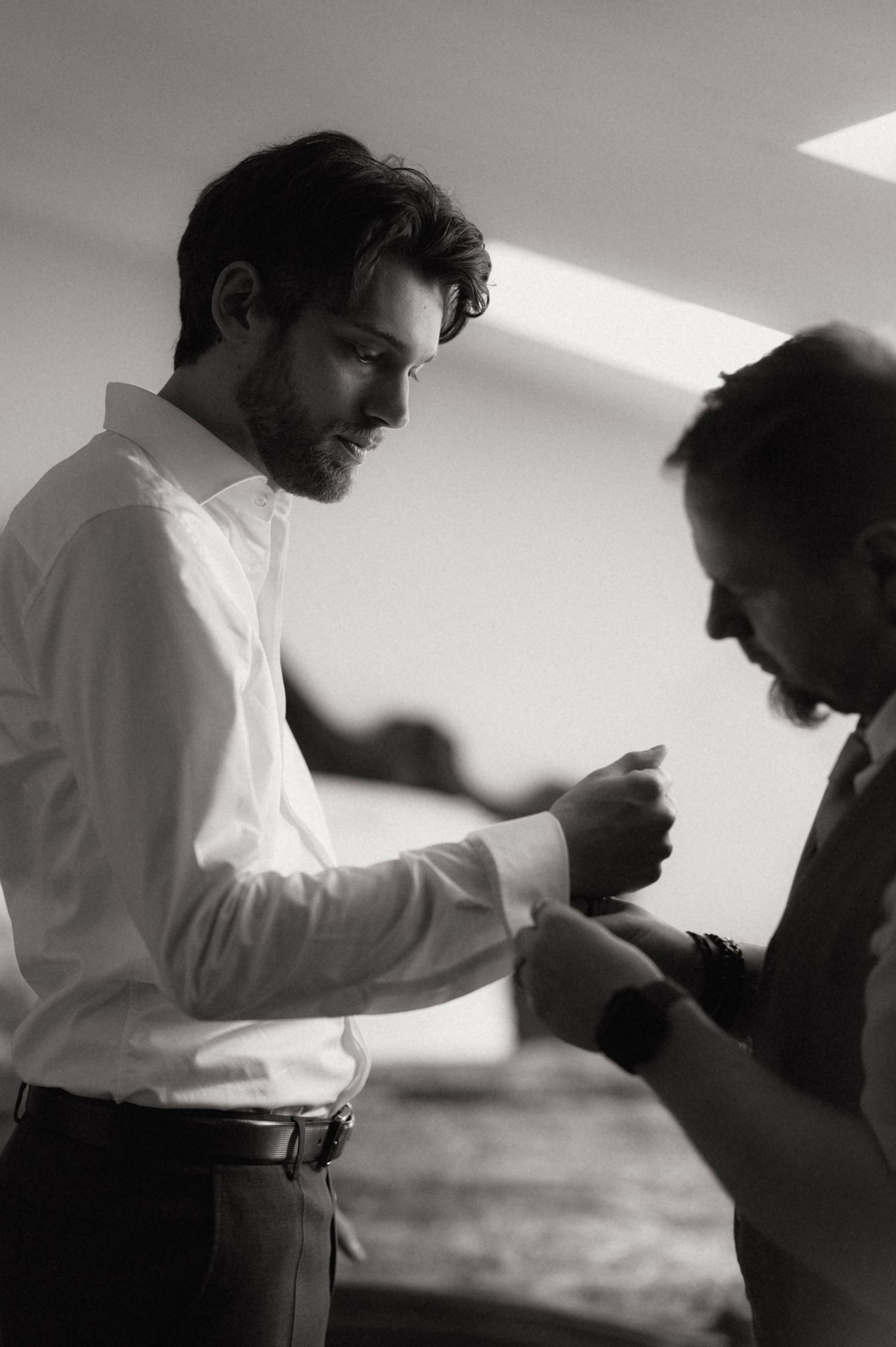 Black and white photo of a groom in a white dress shirt getting his cufflinks fastened by a groomsman.