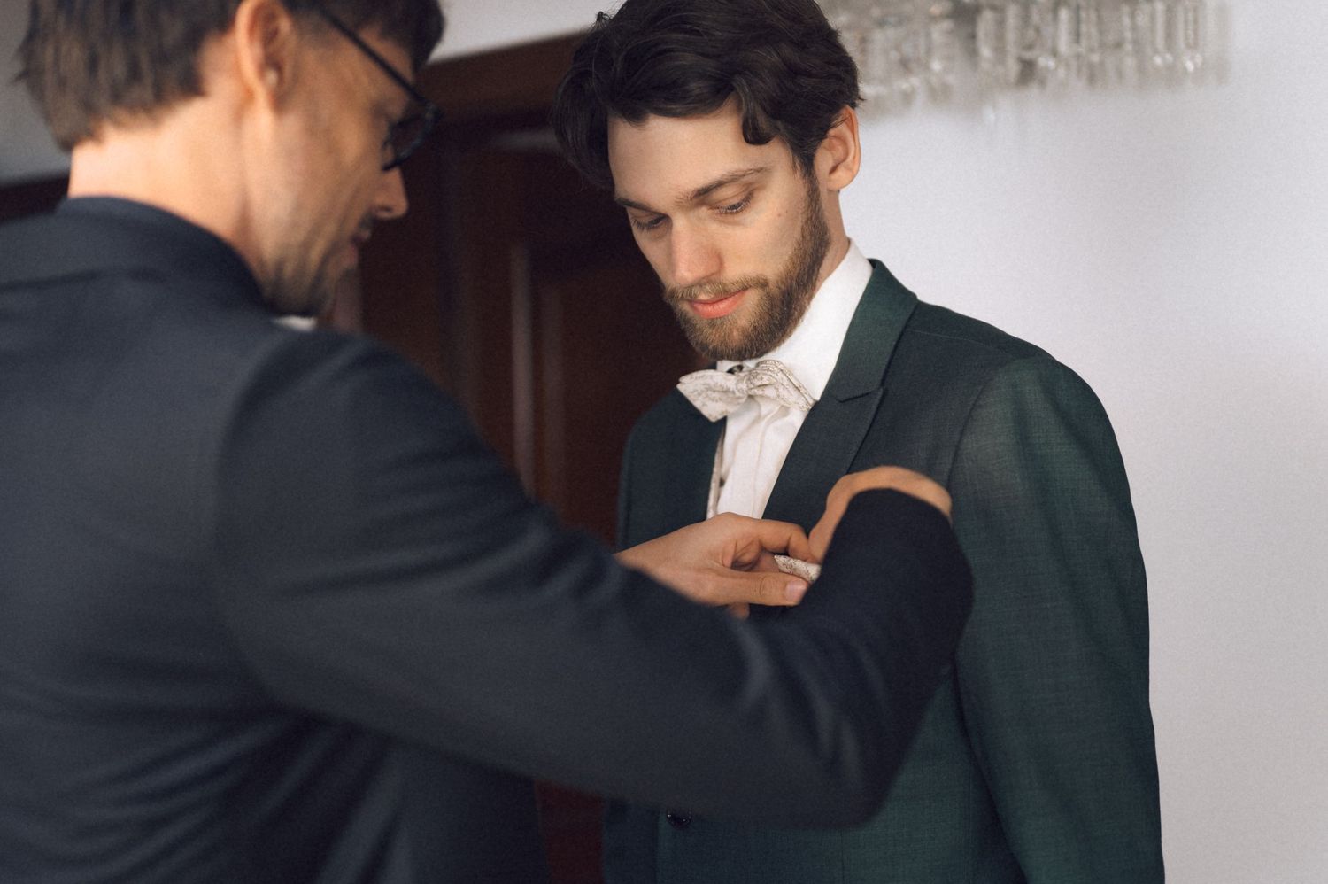 Groomsman helping groom with boutonniere on green suit before wedding.