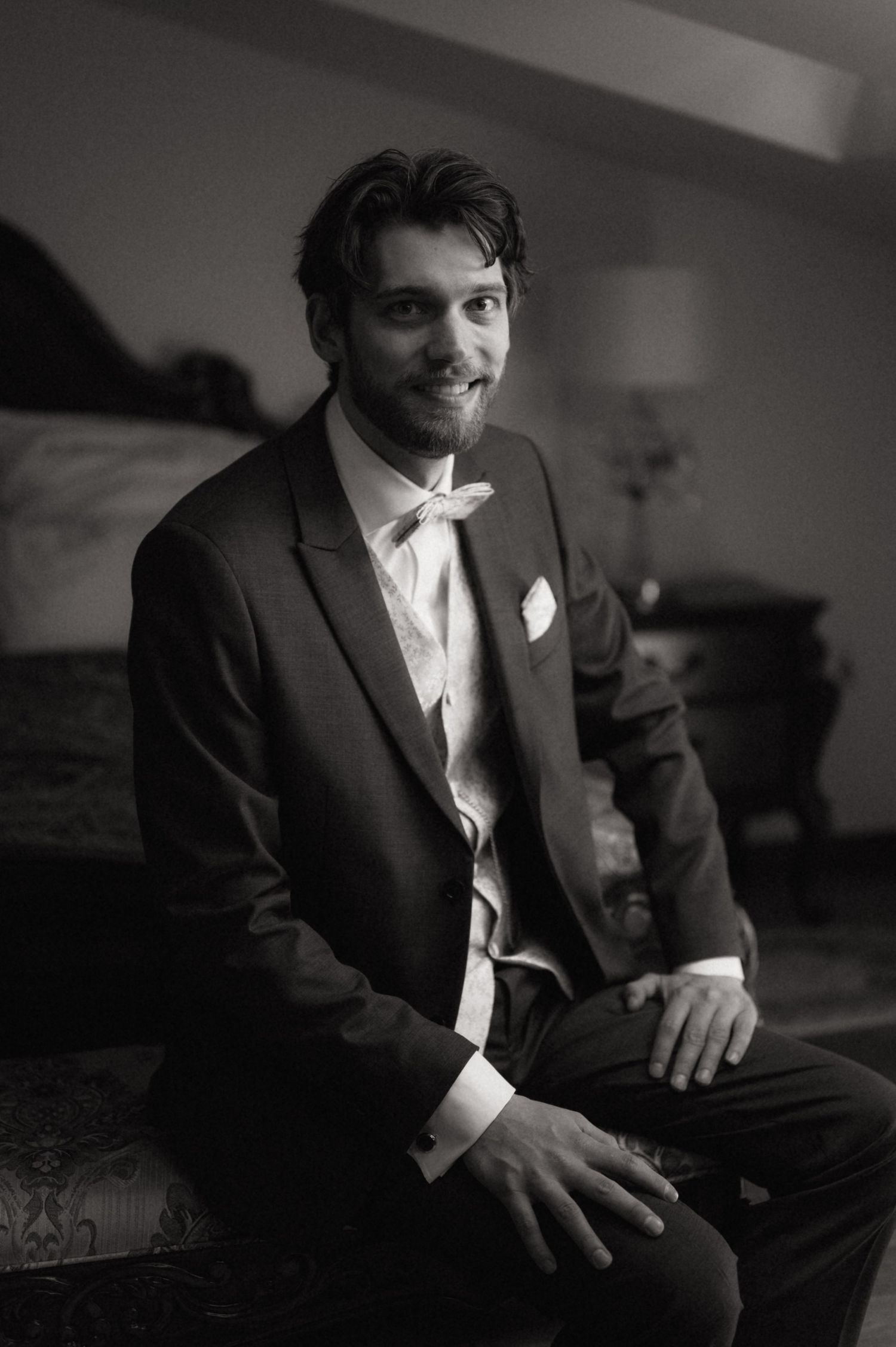 Black and white portrait of a smiling groom in a suit and tie sitting elegantly at a wedding venue.
