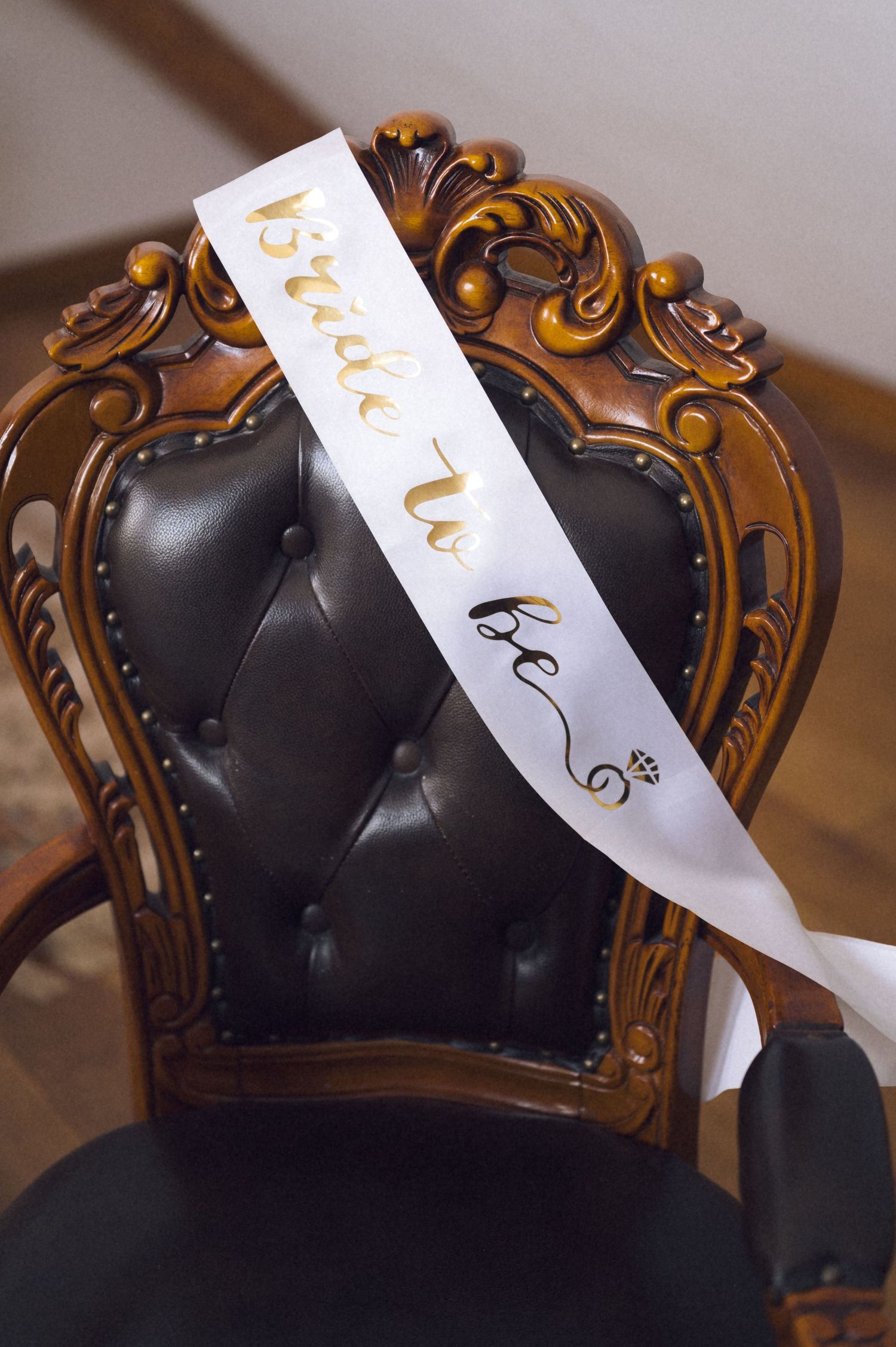 Elegant carved wooden chair with black tufted leather seat displaying a white and gold bride-to-be sash.