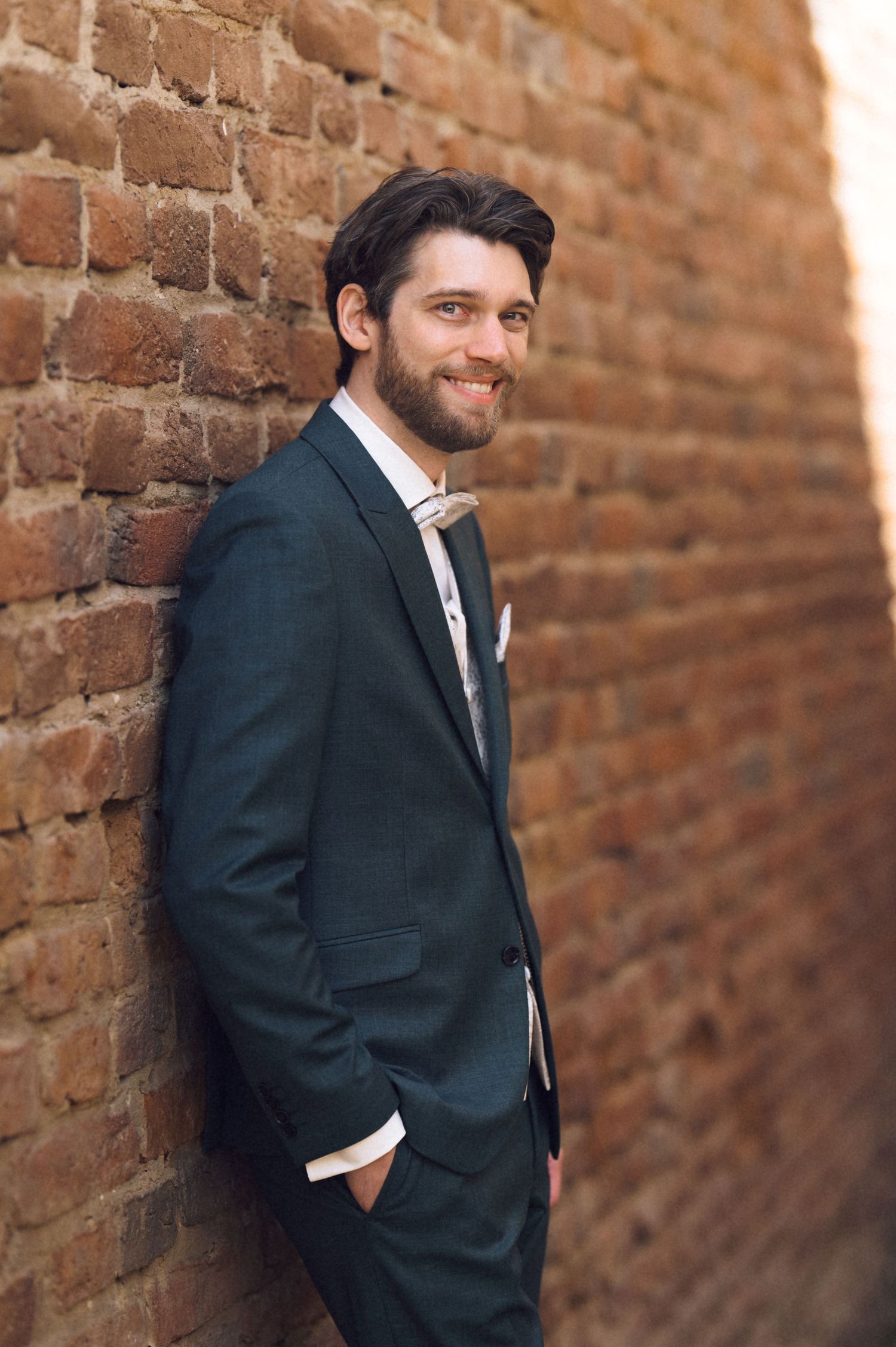 Smiling bearded man in dark teal suit leaning against rustic brick wall, hands in pockets, professional portrait.