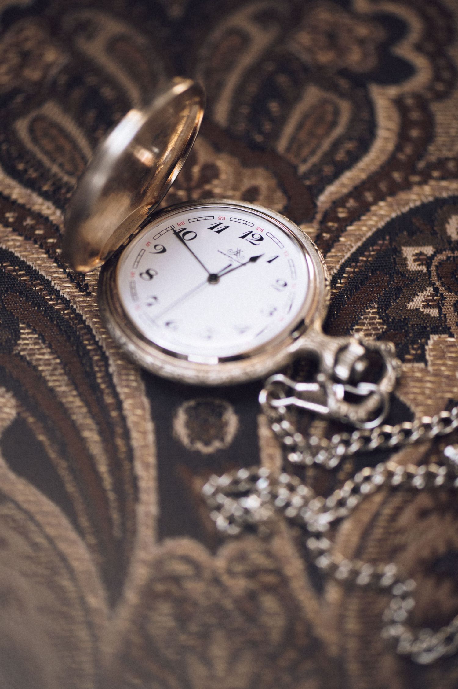 Vintage gold pocket watch with chain on ornate paisley fabric background.