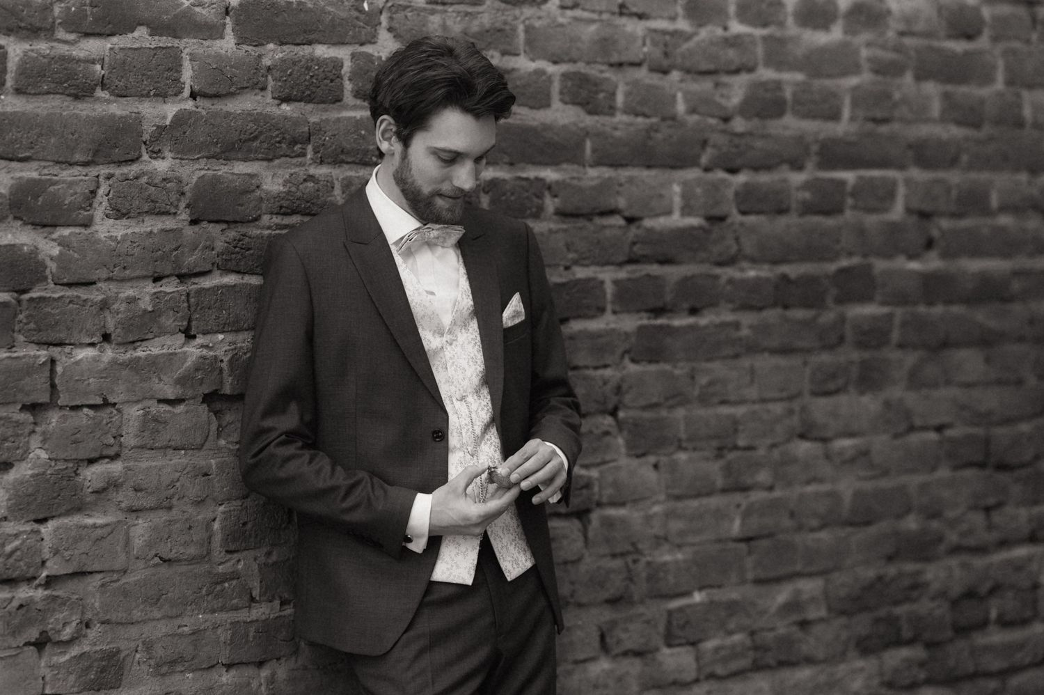 Black and white photo of a bearded man in a suit leaning against a brick wall, looking down thoughtfully.