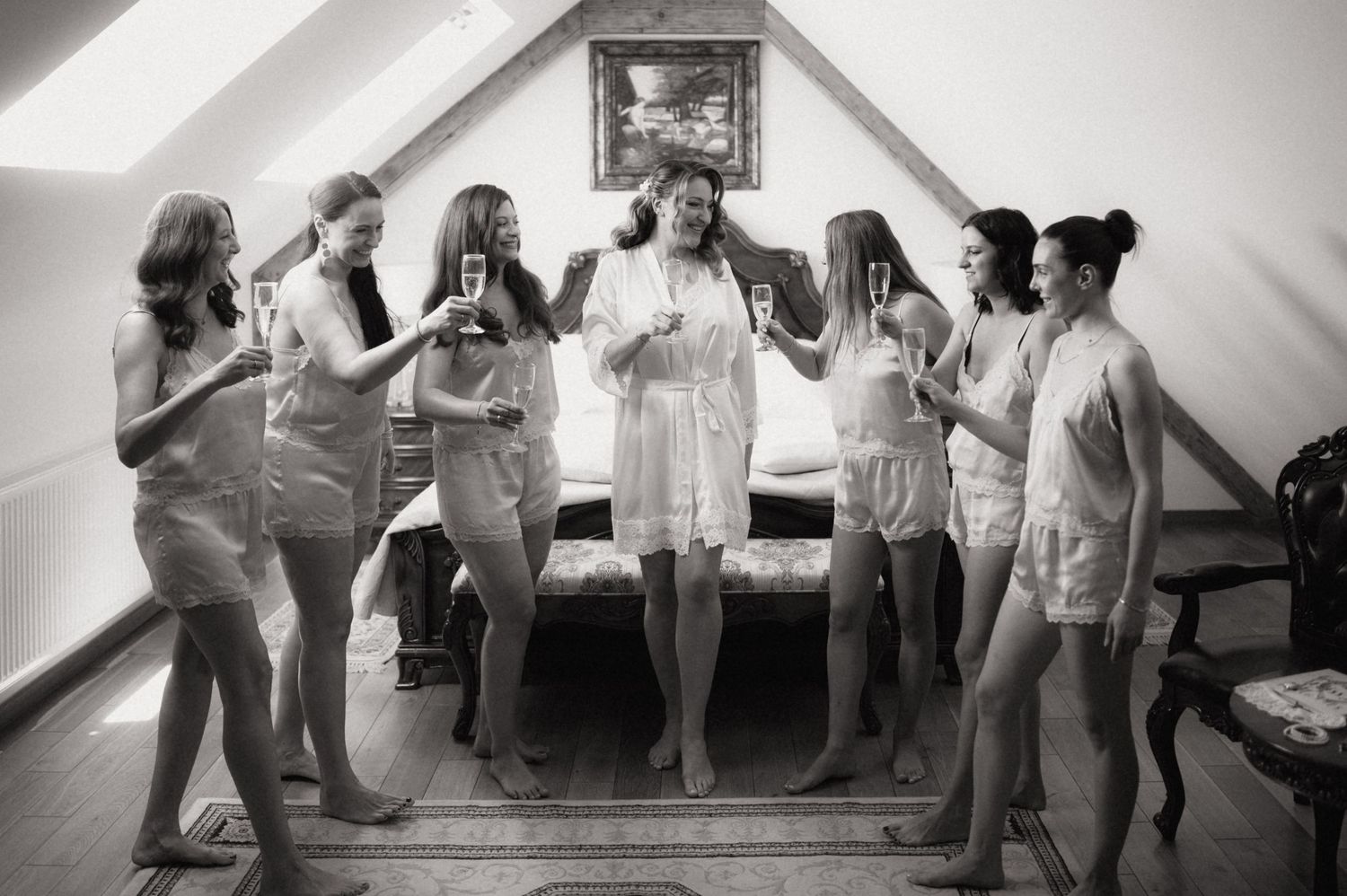 Group of bridesmaids in white pajamas toasting with champagne glasses in a cozy attic bedroom celebration.