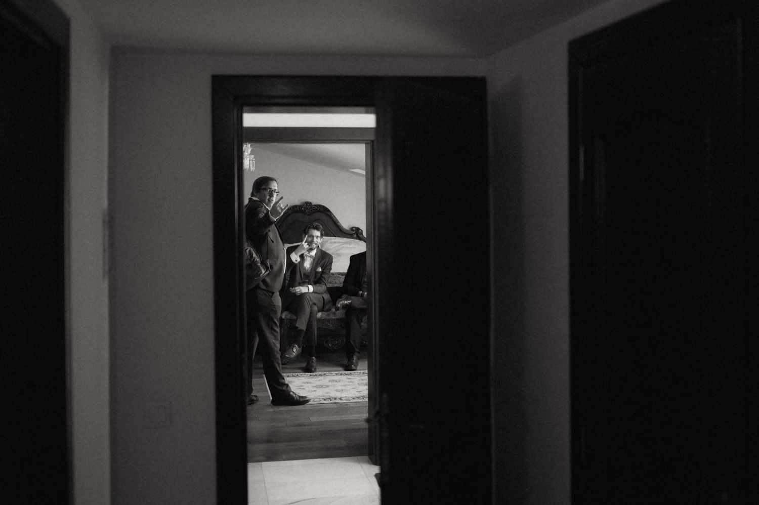 Black and white photo of men in suits preparing in a dimly lit room, viewed through a doorway.