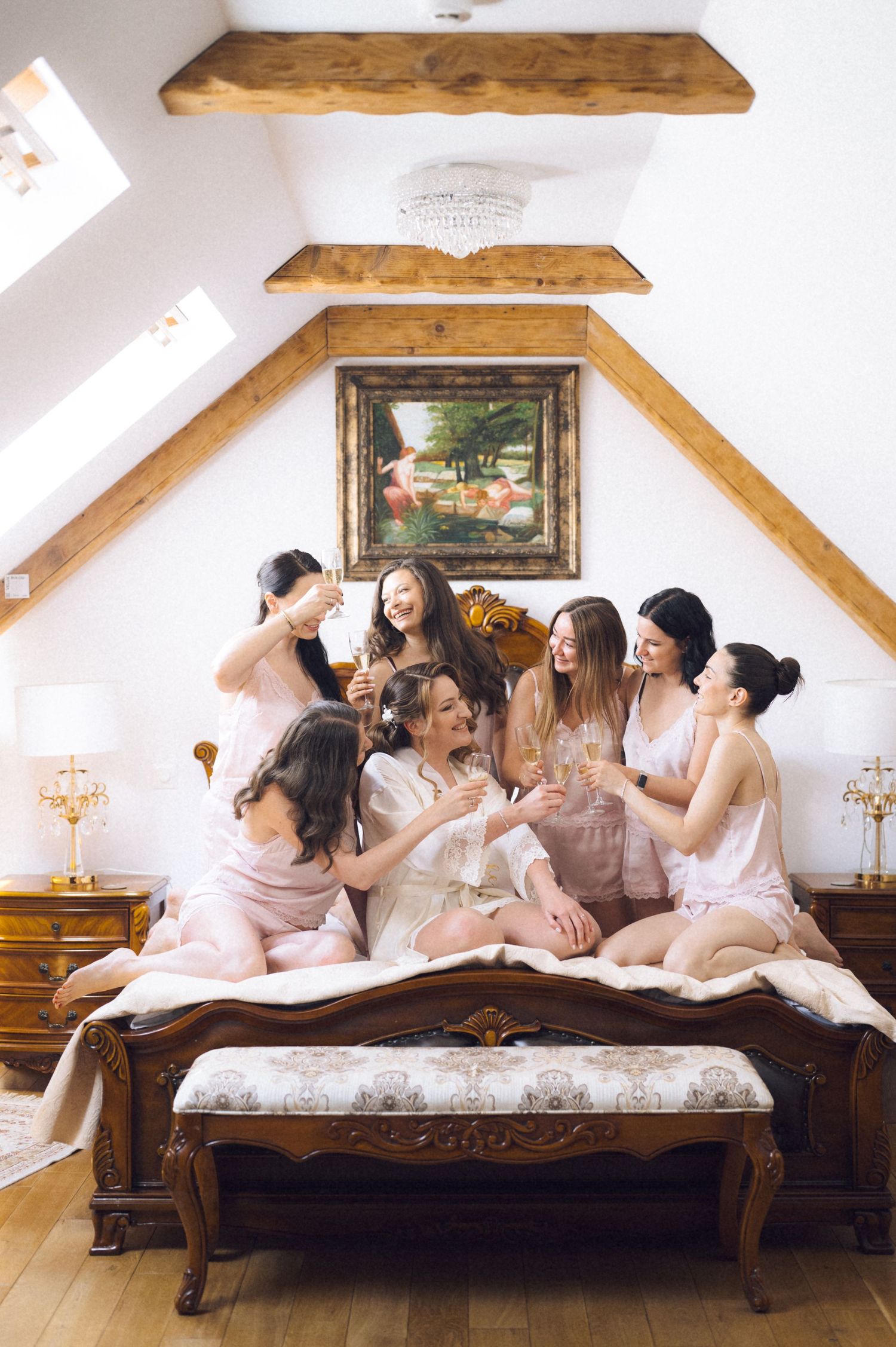 Six bridesmaids in pink satin pajamas toasting champagne on an ornate wooden bed in a charming attic bridal suite.