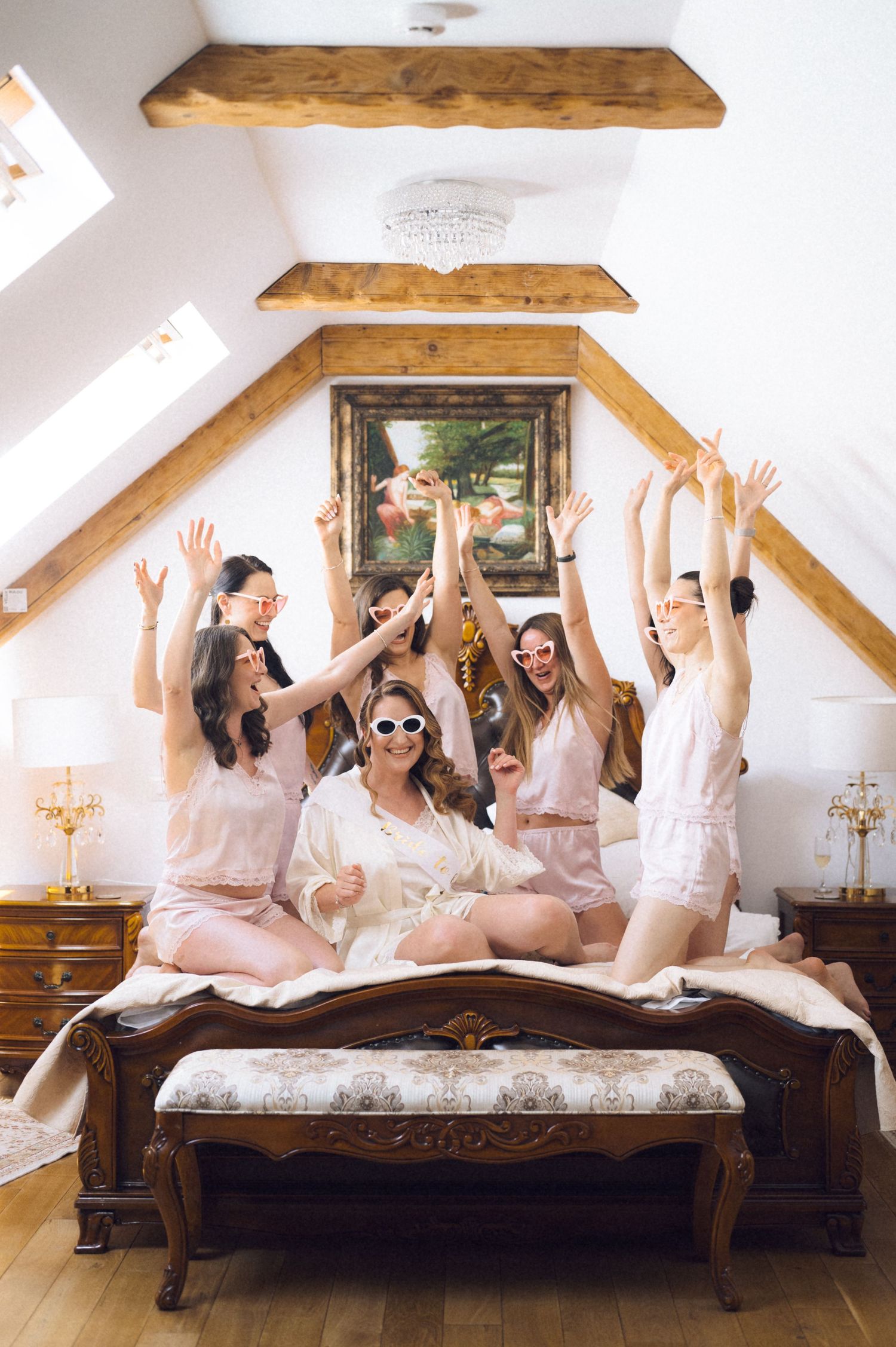 Group of bridesmaids in pink pajamas celebrating on ornate bed in rustic attic bedroom with raised arms.