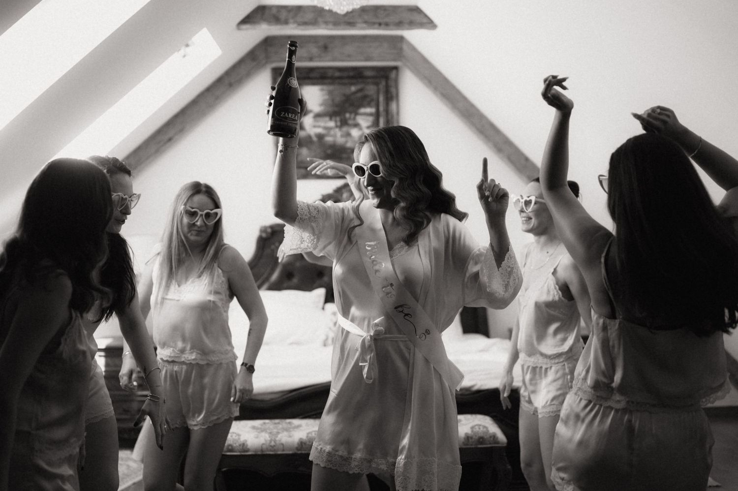 Black and white photo of a bridal party celebrating in robes, dancing and posing in an attic bedroom.