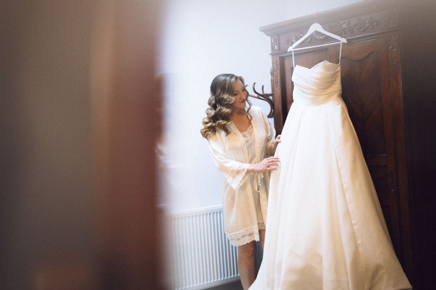 Bride in white robe admiring her elegant strapless wedding gown hanging on a wooden wardrobe door.