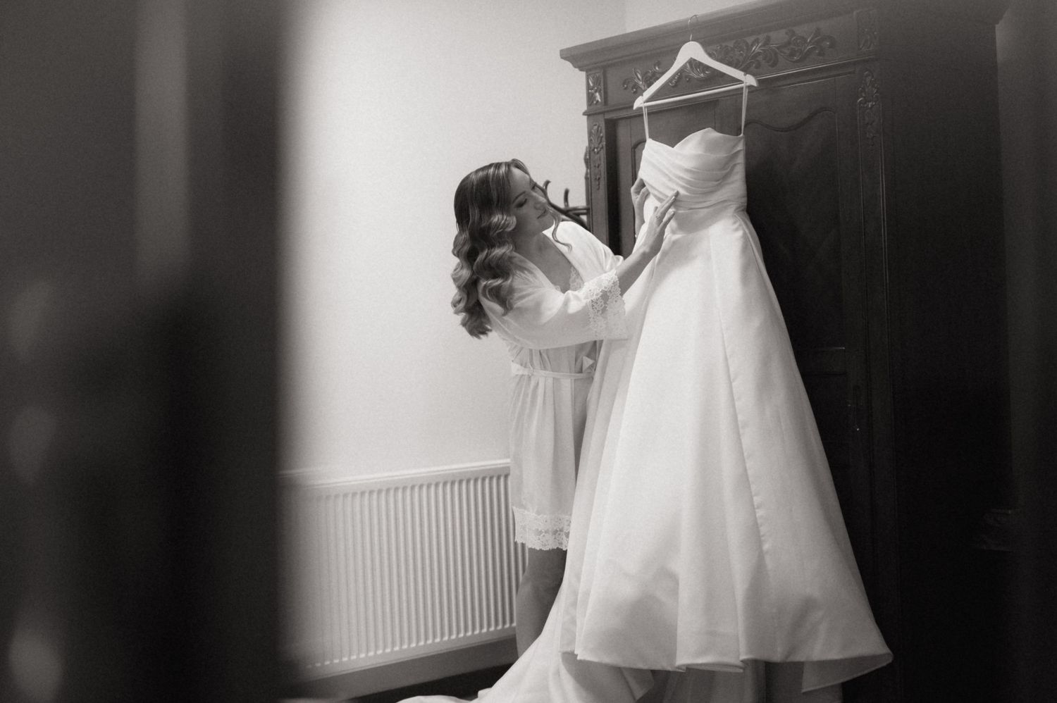 Black and white photo of a bride admiring her wedding dress hanging on a wardrobe door.