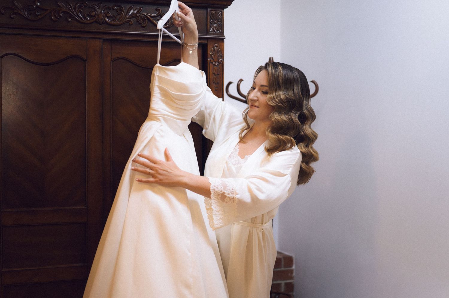 Bride holding up elegant white wedding dress in front of wooden armoire while wearing a white robe.
