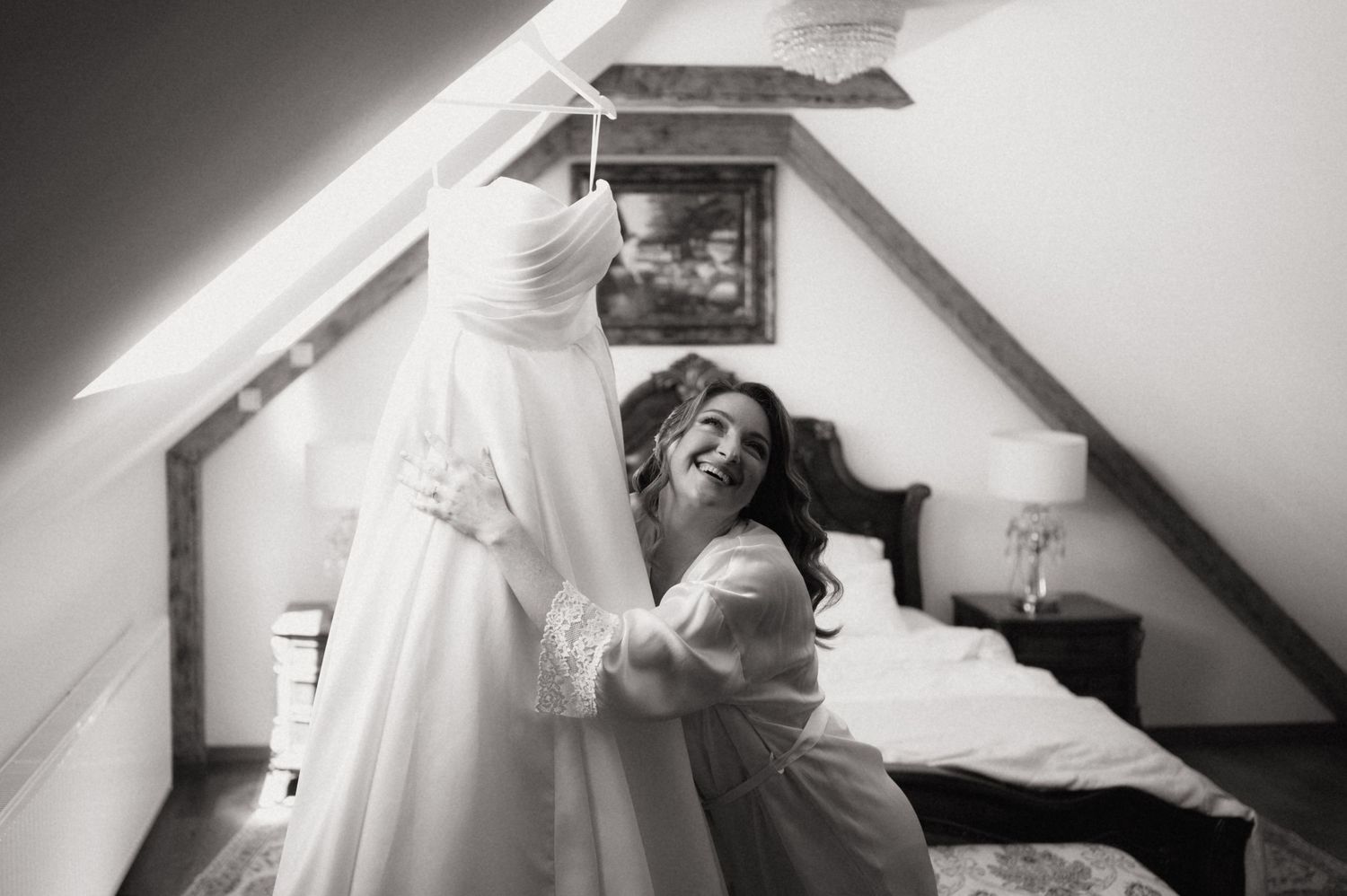 Bride laughing joyfully in robe while holding wedding dress in attic bedroom, black and white photo.