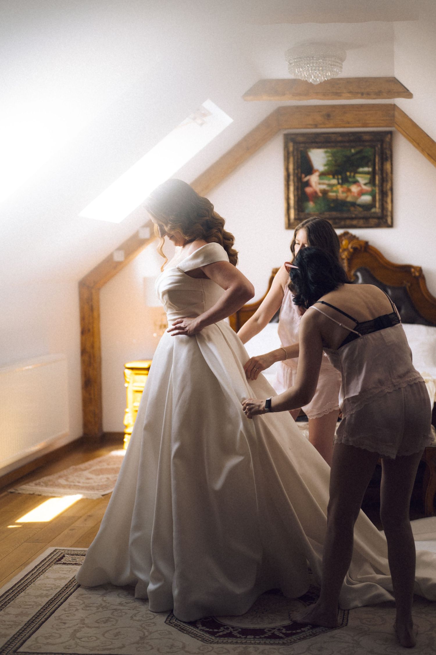 Bride being helped into elegant white ball gown wedding dress by bridesmaid in sunlit attic bridal suite.