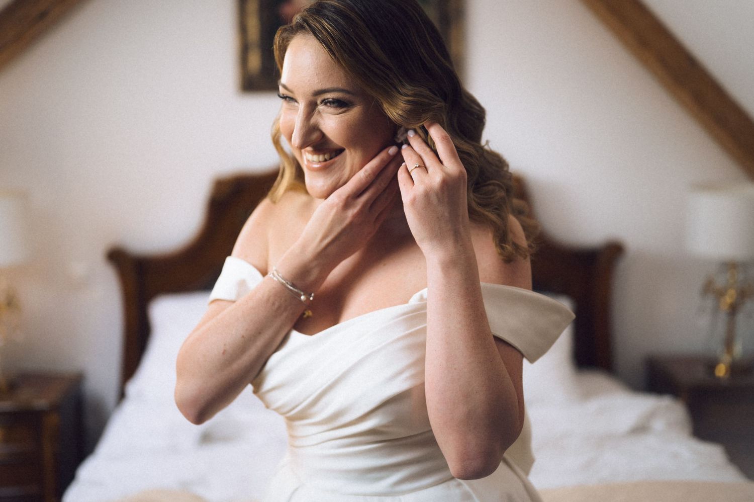 Bride in white off-shoulder gown putting on earrings while sitting on bed during wedding preparations.