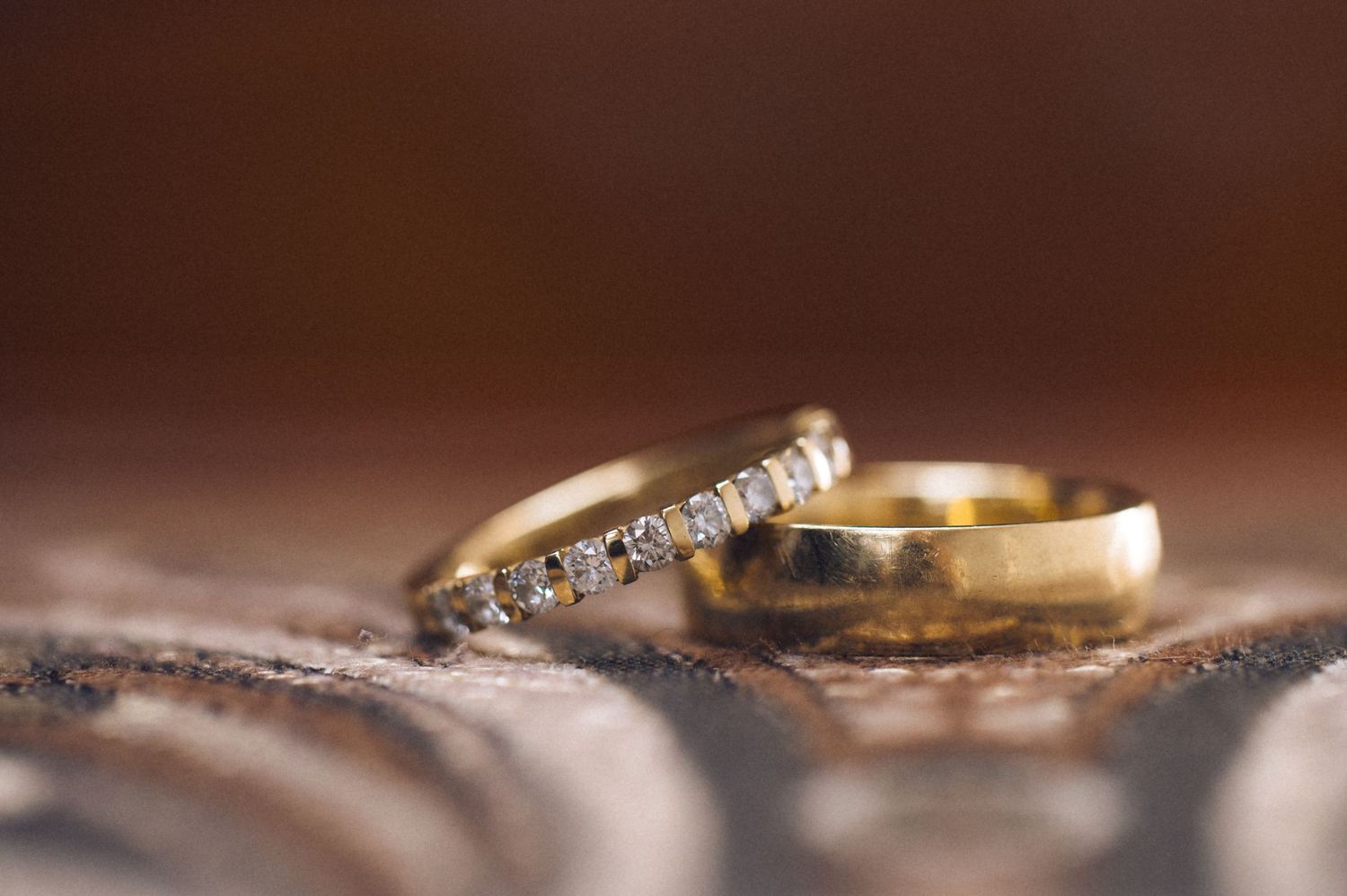 Two gold wedding rings, one with diamond accents, resting on a textured surface with a warm brown bokeh background.