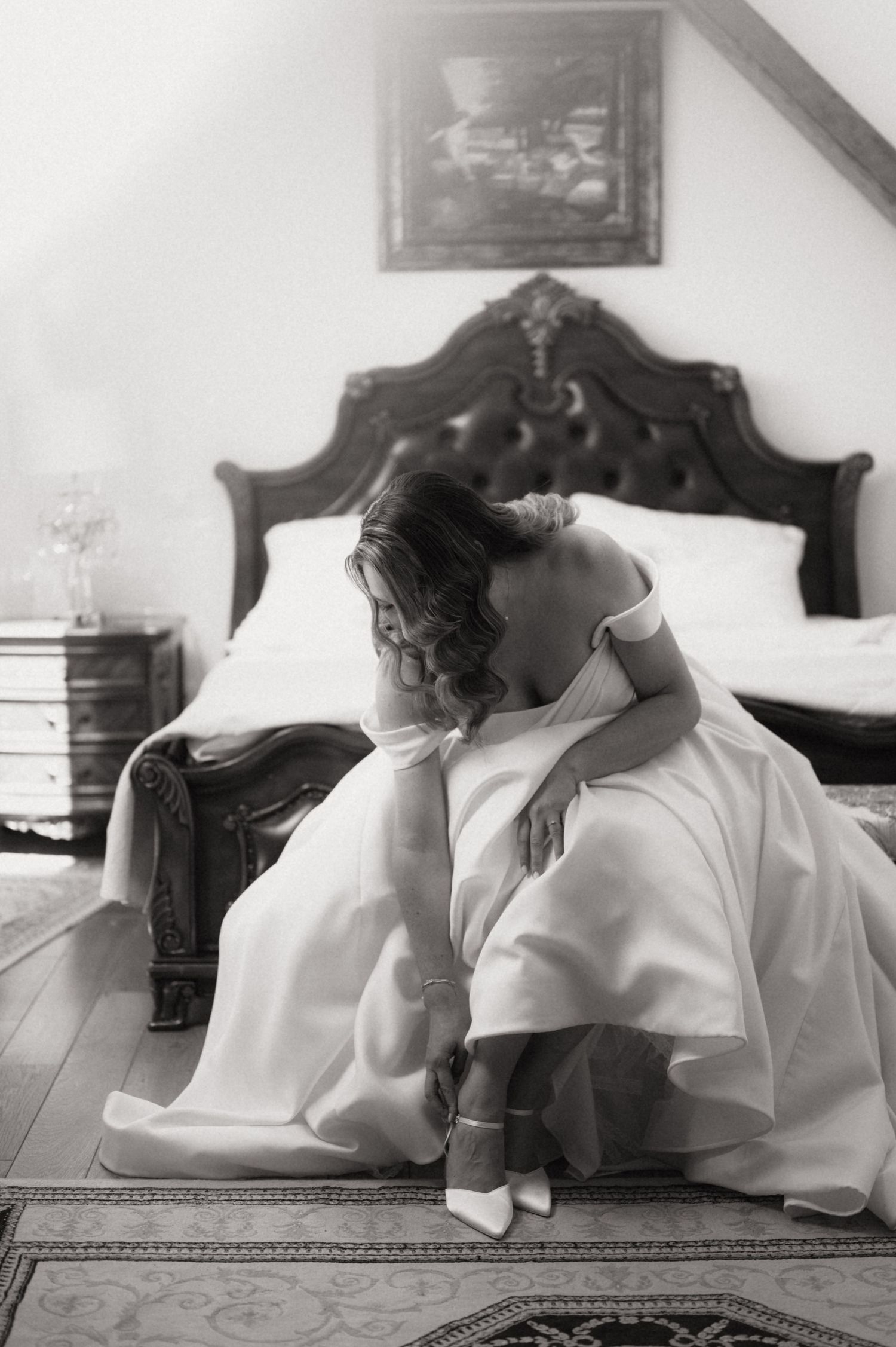 Black and white photo of a bride in a white gown sitting on bedroom floor, adjusting her heels near an ornate bed.