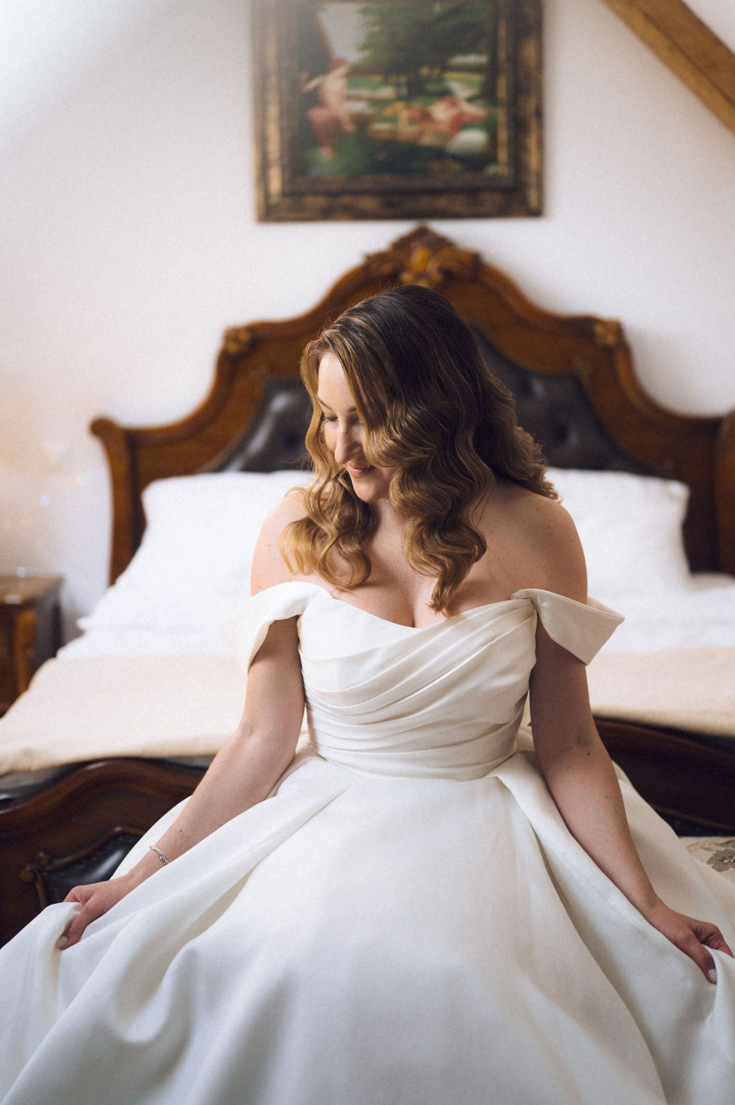 Bride in off-shoulder white ball gown sitting on bed with ornate wooden headboard, looking down gracefully.