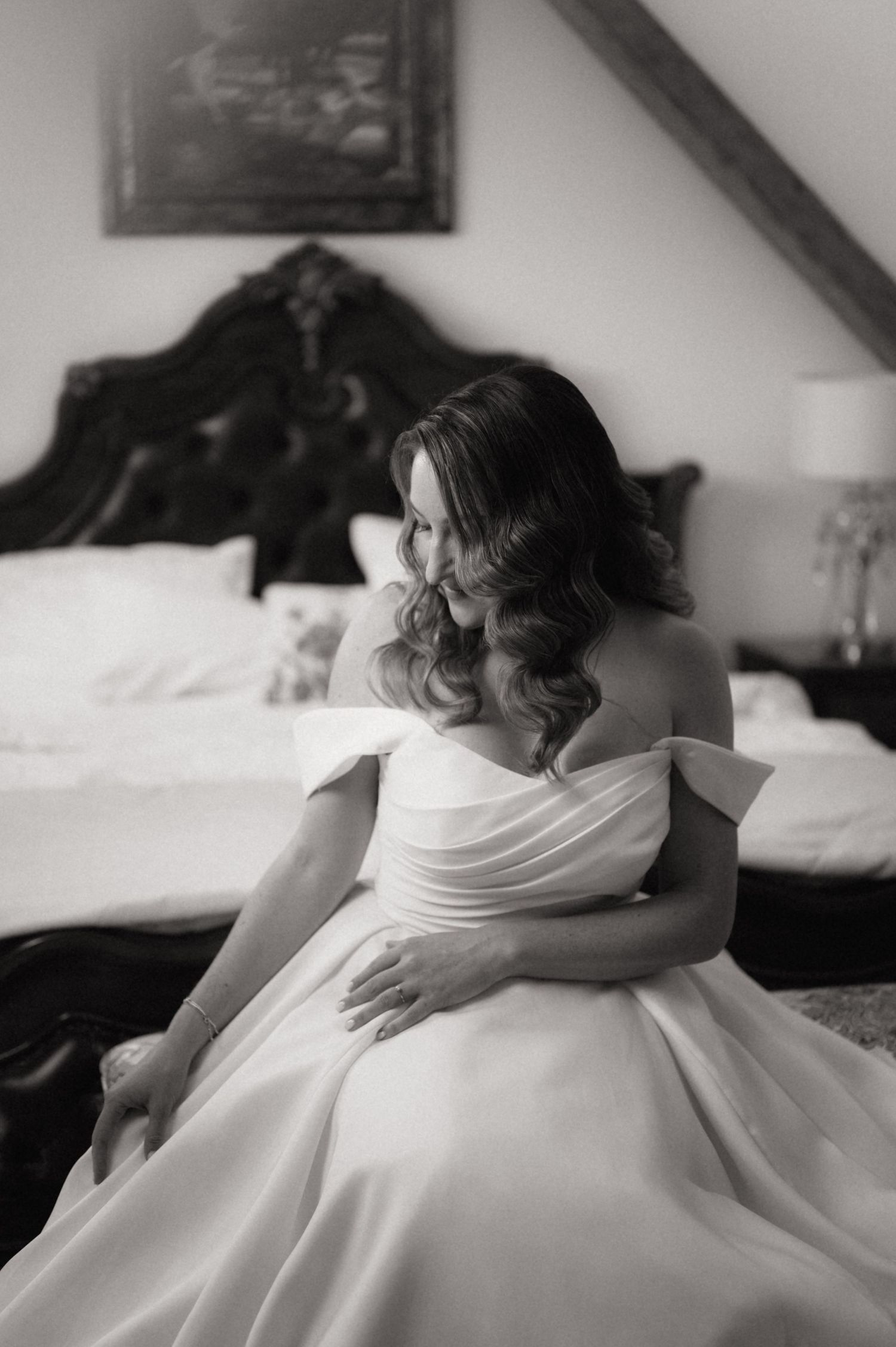 Black and white photo of a bride in an off-shoulder wedding gown sitting elegantly in a bedroom.