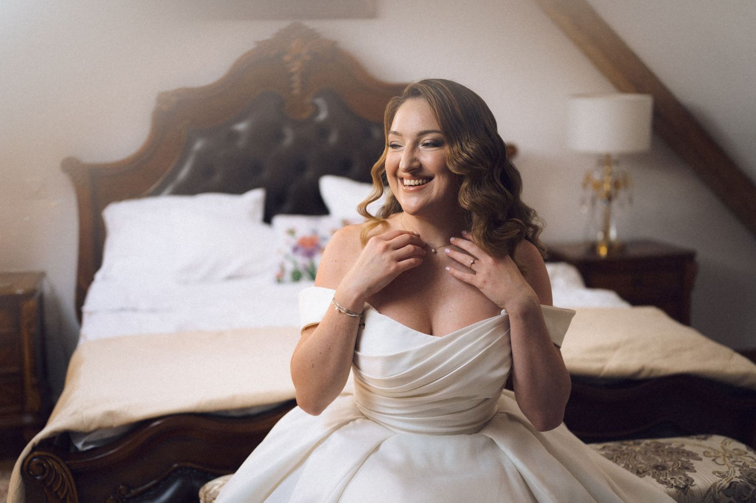 Smiling bride in strapless white wedding gown sitting on bed in elegant room with wooden headboard.