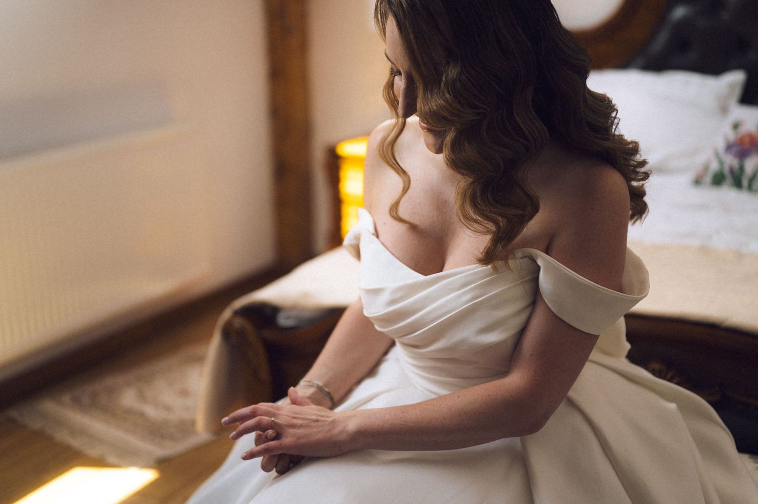 Bride in elegant off-shoulder white wedding gown sitting gracefully, showcasing beautiful back detail and flowing skirt.