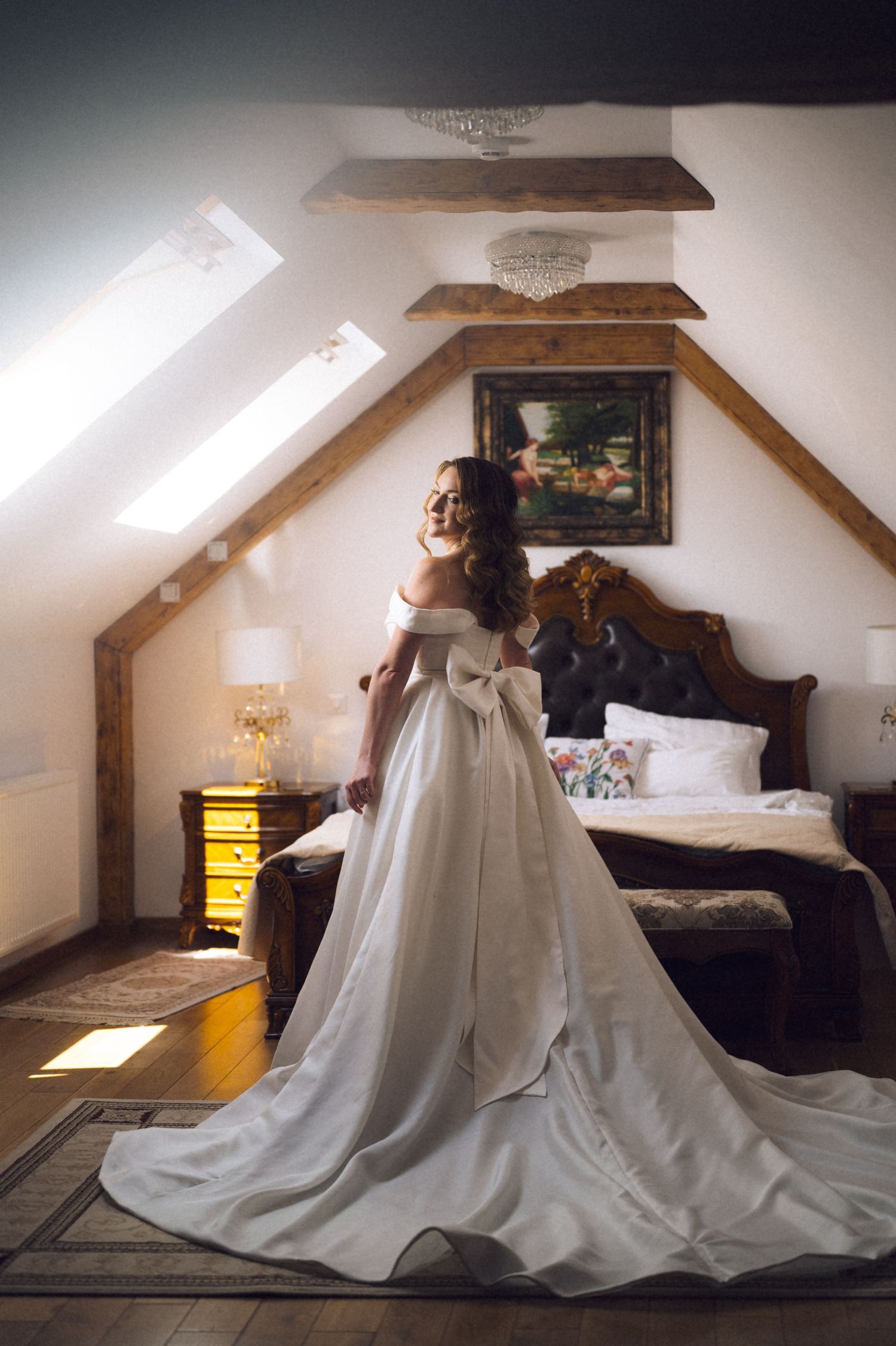 Bride in elegant off-shoulder white gown with long train poses in rustic attic bedroom with wooden beams.