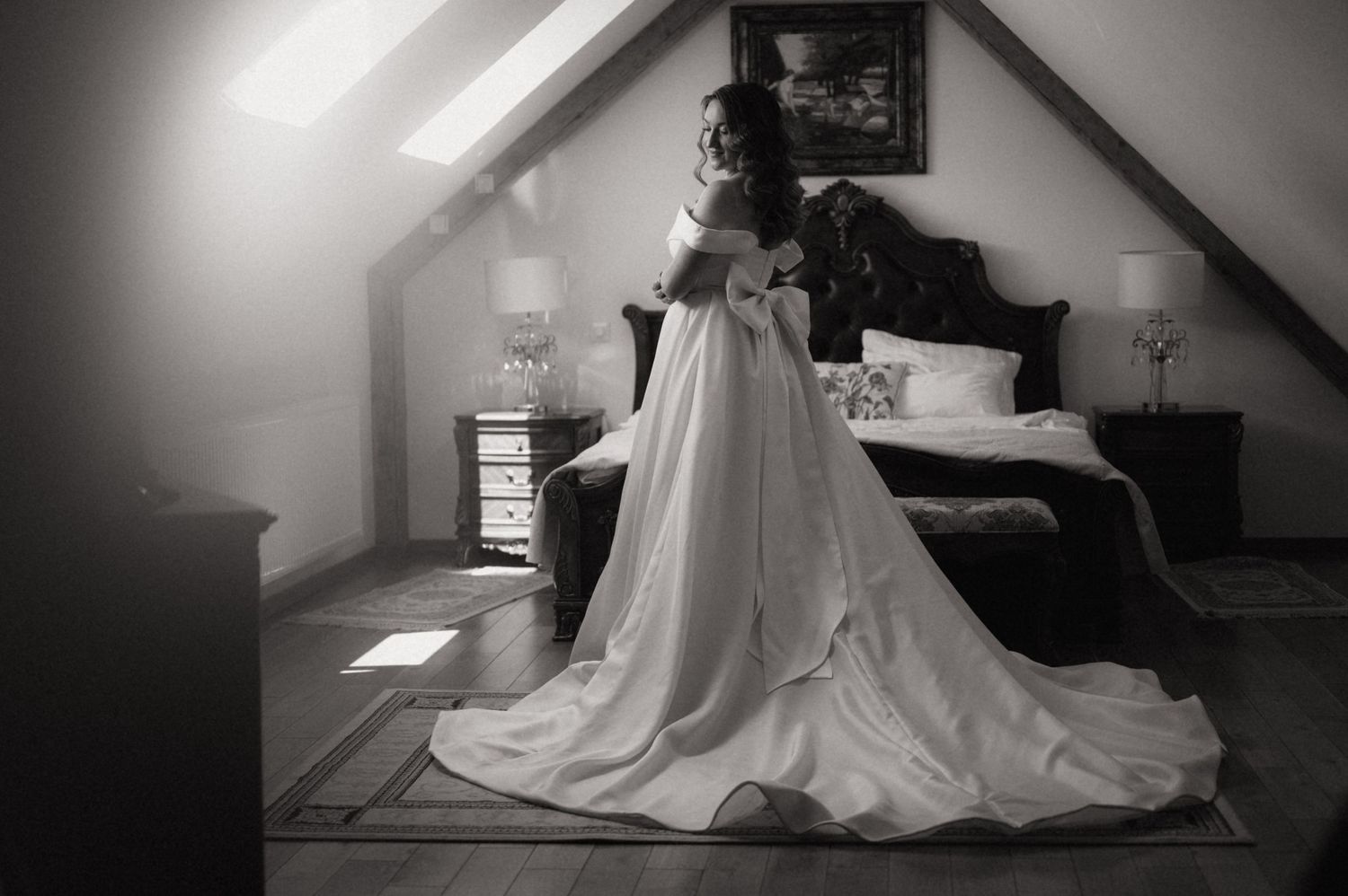 Black and white photo of a bride in a flowing wedding gown standing by a bedroom window in an attic room.