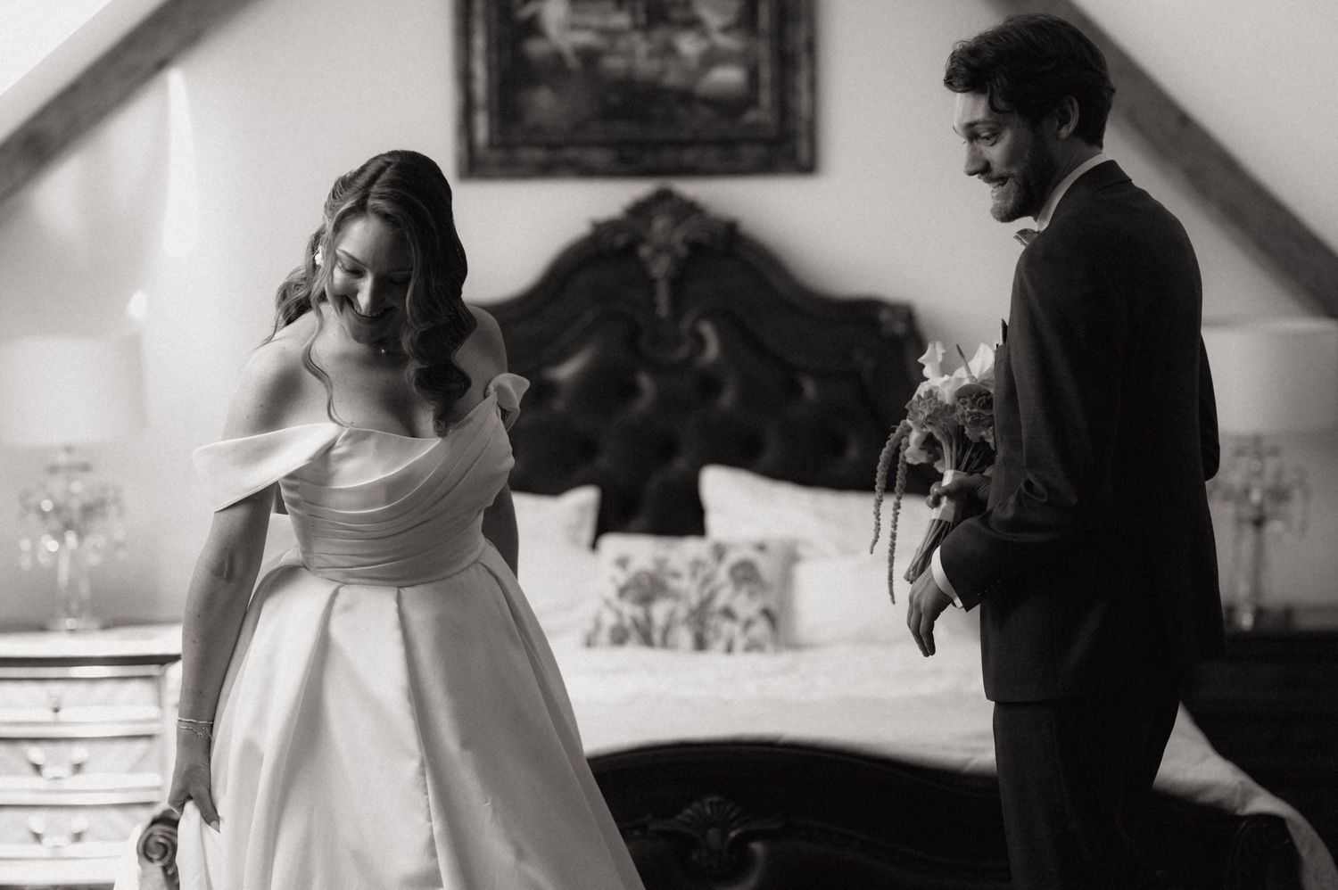 Black and white wedding photo of bride in off-shoulder gown and groom holding bouquet in elegant bedroom.