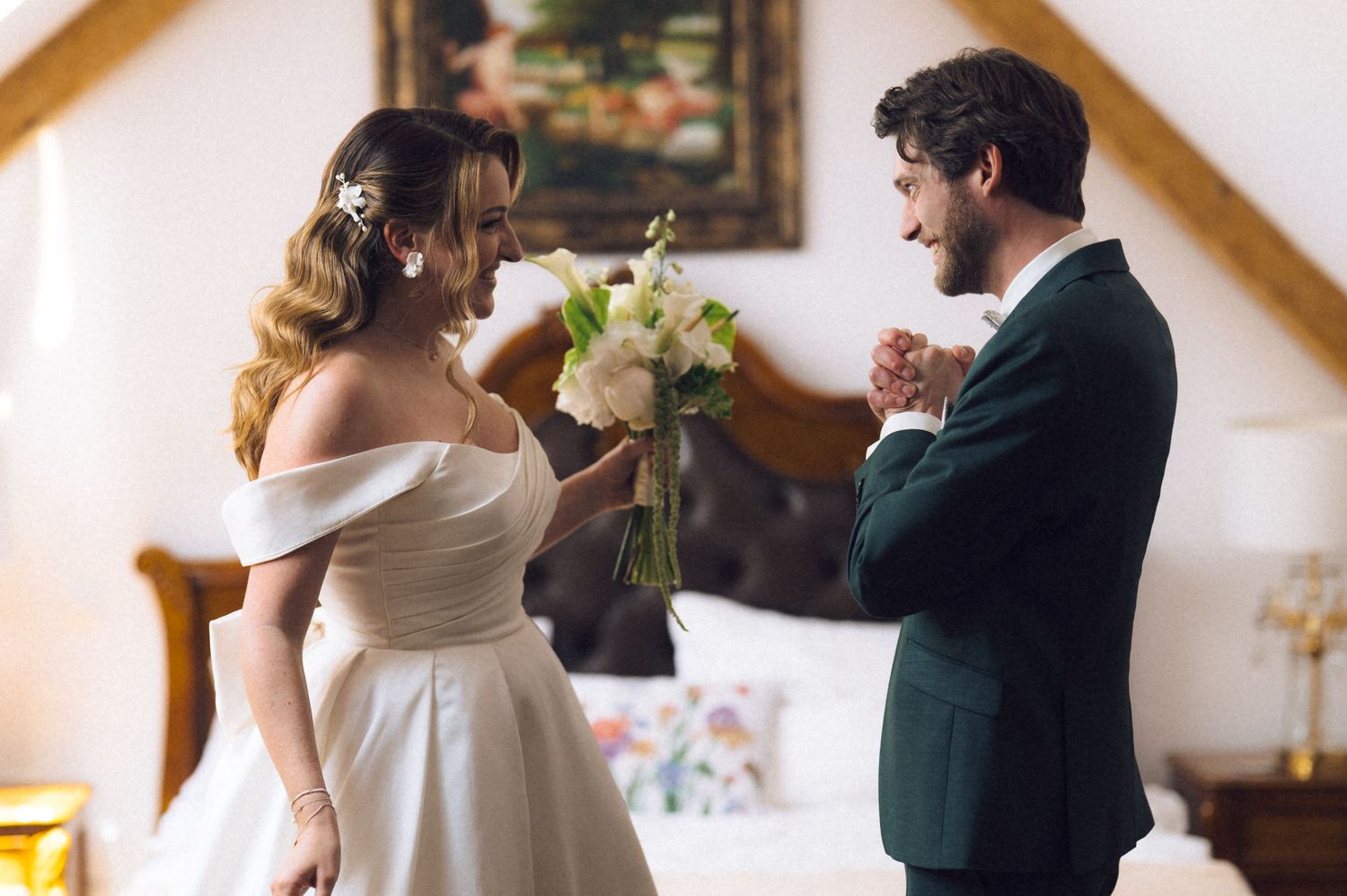 Bride in white off-shoulder gown holds bouquet while groom in dark suit admires her in elegant room.