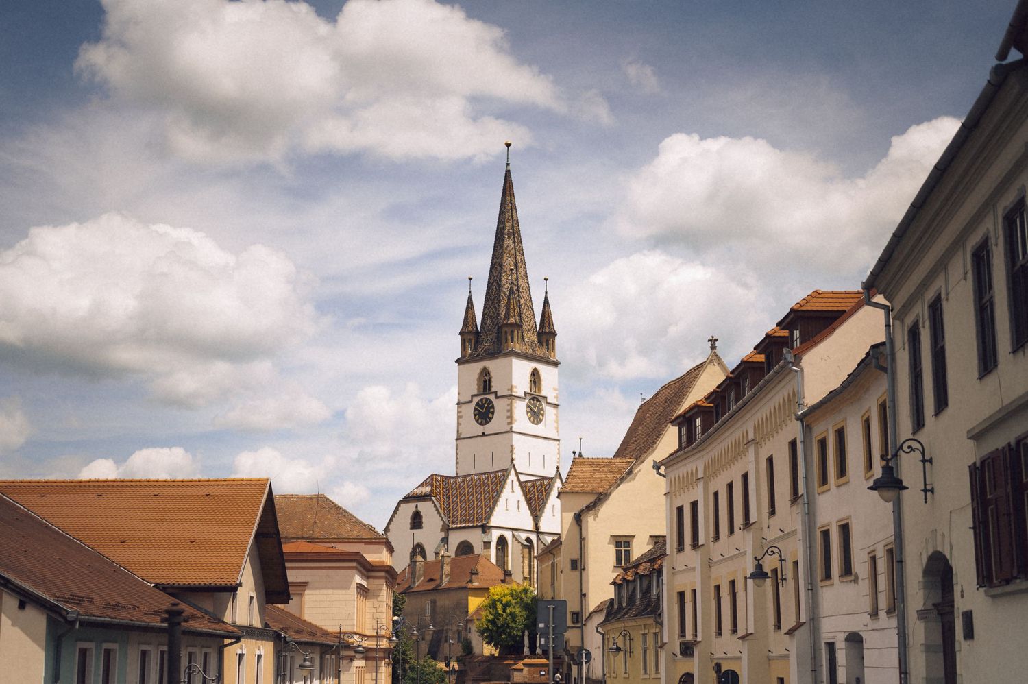Historic European town street with a tall Gothic church spire rising above colorful buildings under a cloudy sky.