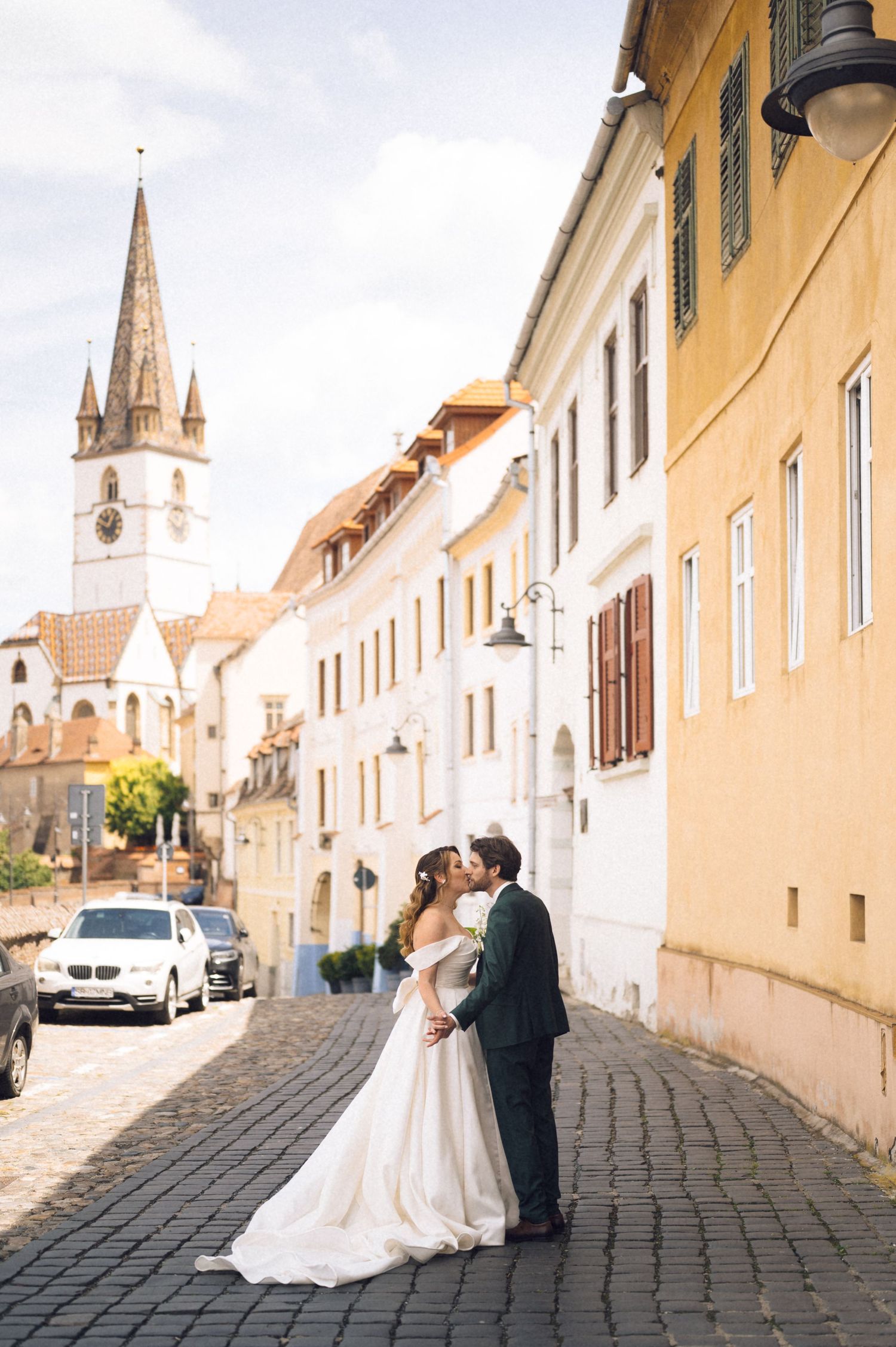 Bride and groom kissing on cobblestone street in Sibiu, Romania, with historic church tower and colorful buildings in background.