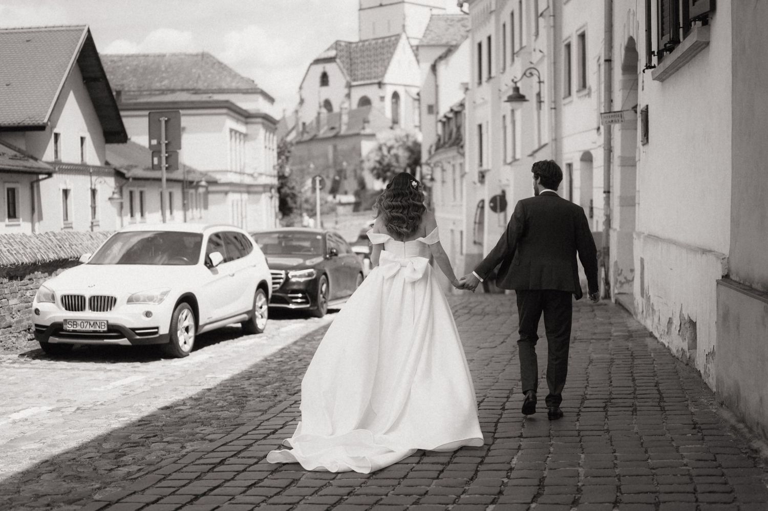 Black and white photo of a bride and groom walking hand in hand down a cobblestone street past a white BMW.